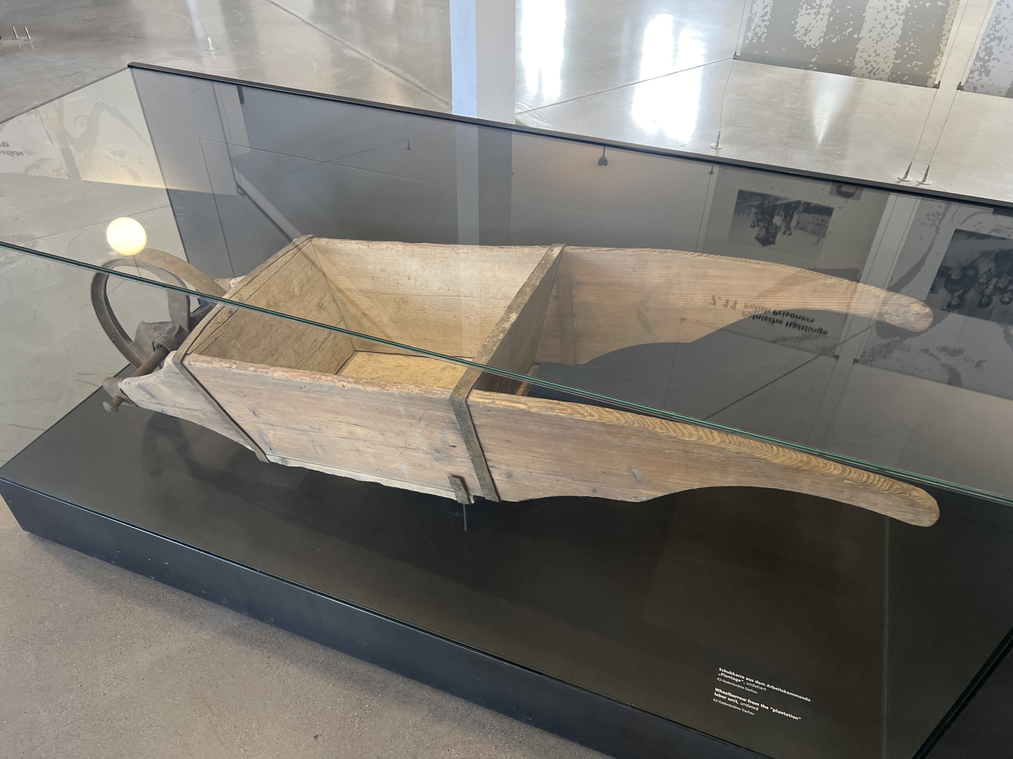 An exhibit, possibly in the Dachau Concentration Camp Memorial Site, features a wooden wheelbarrow displayed in a glass case. The wheelbarrow is weathered and has two compartments. A metal wheel is attached to the front, and handles extend from the back. A lightbulb is visible above the wheel. In the background, enlarged black and white photographs are displayed on a wall. A descriptive plaque is affixed to the interior of the glass case, providing information about the wheelbarrow and its origin, with text in both German and English, identifying it as a "Wheelbarrow from the 'plantation'" and mentioning it is from the "labor unit, undated" at the "KZ-Gedenkstätte Dachau". The flooring of the exhibit space appears to be polished concrete.