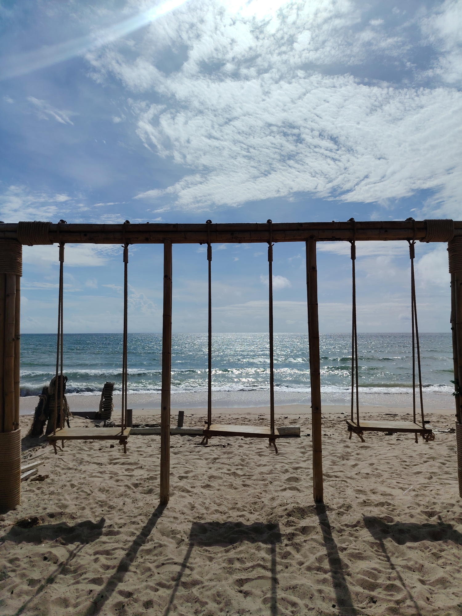This is a forensic, matter-of-fact description of an image depicting a beach scene. The location is identified as Choeng Thale, Thailand.
The image shows a wooden swing set constructed from bamboo poles and ropes, positioned on a sandy beach facing the ocean. The swing set has a horizontal bamboo beam supported by vertical bamboo poles. There are multiple swings hanging from the beam, made of wooden planks. Some swings appear to be in use, evidenced by their tilted positions, while others are stationary.
The beach is covered in light-colored sand with footprints and shadows cast by the swing set and possibly other unseen objects. In the background, the ocean stretches to the horizon, with gentle waves breaking on the shore. The water is a clear blue and reflects the sunlight, creating a shimmering effect. The sky above is a bright blue, dotted with scattered white clouds. A sunbeam is visible in the upper left portion of the frame, indicating a bright, sunny day.
To the left of the swing set, there are some debris or remnants of older structures made of wood and possibly palm fronds. These objects are partially buried in the sand. No people are visible in the image. The overall environment suggests a tranquil and scenic beach location.