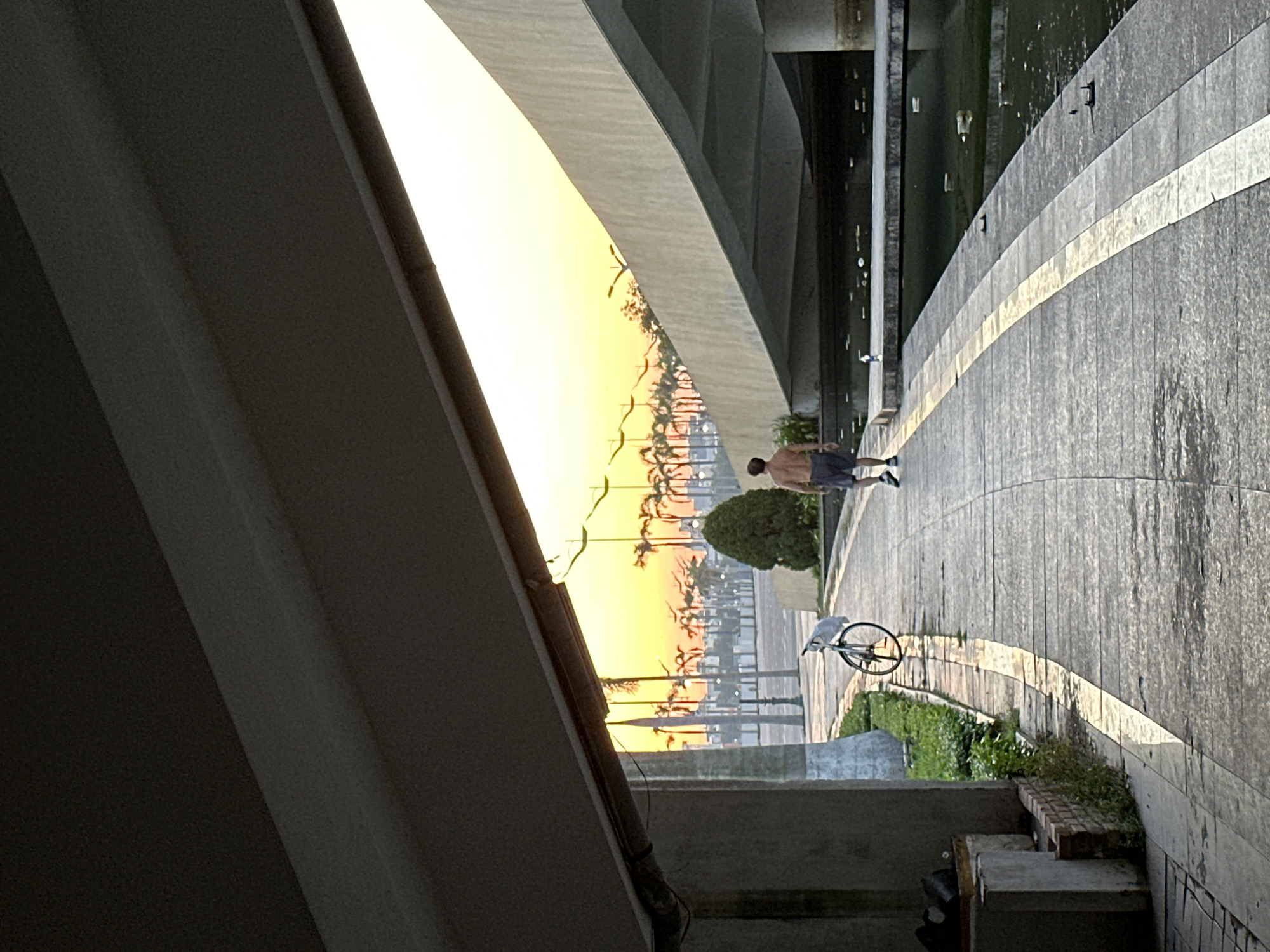 The image depicts a person walking on a paved pathway under a bridge in Da Nang, Vietnam. The pathway is bordered by a water channel on the right and greenery on the left. In the distance, palm trees are silhouetted against a vibrant orange and yellow sunrise sky. A bicycle is parked on the left side of the pathway. The scene is characterized by the warm glow of the sunrise and the geometric lines of the bridge and pathway.