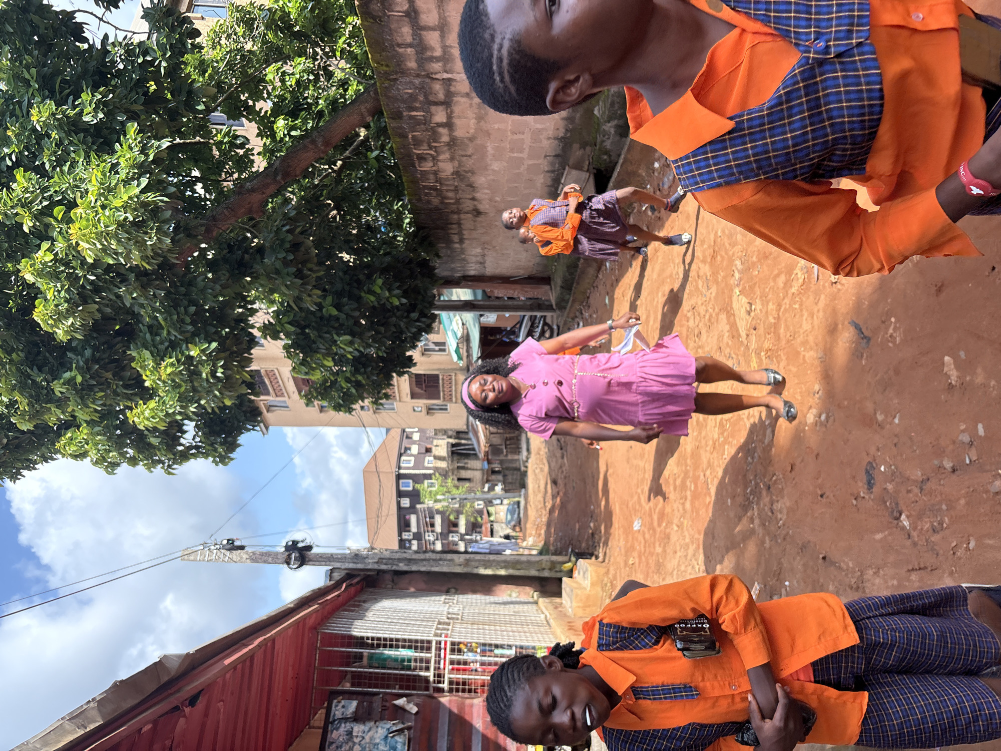 The image shows a group of children, likely students, dressed in school uniforms. The uniforms consist of orange long-sleeved shirts with blue and yellow checkered collars and blue checkered skirts. One student in the foreground is wearing a blue and yellow checkered skirt and an orange shirt with a checkered collar. Another child in the mid-ground is wearing a similar uniform and appears to be walking away from the camera. In the background, two more children in matching uniforms are visible.
To the left of the frame, a large tree with green foliage dominates the upper portion. Behind the tree, buildings with visible windows and balconies can be seen. A power line with a pole runs across the sky. The ground appears to be made of reddish-brown earth, suggesting a rural or semi-urban setting. The lighting indicates it is daytime, with the sun casting shadows on the ground. The overall mood seems casual and everyday, with the children likely on their way to or from school, or perhaps during a break.
One of the cropped images shows a close-up of the shirt worn by the child in the foreground. On the shirt, there is a label that reads "OXFORD". The context of Nkpor, Nigeria, suggests this is a street scene in that region.