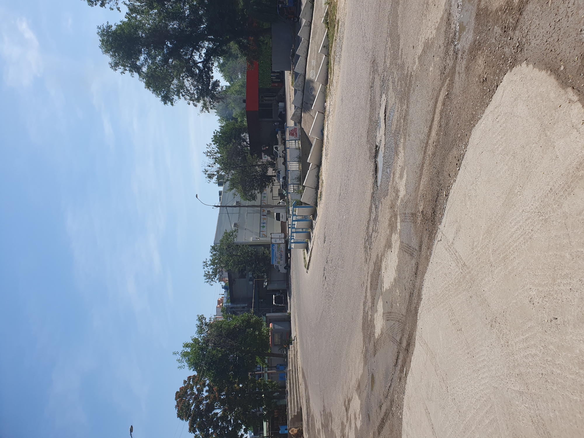The image depicts a view of a street corner with a light blue sky overhead. In the foreground, a paved road shows signs of wear and tear, with puddles and tire tracks visible. To the left, a concrete structure with steps is present, alongside some stacked lumber and a blue post. Beyond the road, several trees with green and reddish foliage are scattered throughout the scene.
In the middle ground, a white truck with a logo on its side is parked. There are also other vehicles, including a white pickup truck and a blue vehicle. Buildings, appearing to be industrial or commercial in nature, line the background. One building has a red awning. A street lamp is visible on the left side of the image.
The scene is suggestive of an urban or industrial area in Tashkent, Uzbekistan, given the architecture and vegetation. The overall impression is one of an ordinary day in a functional part of the city.