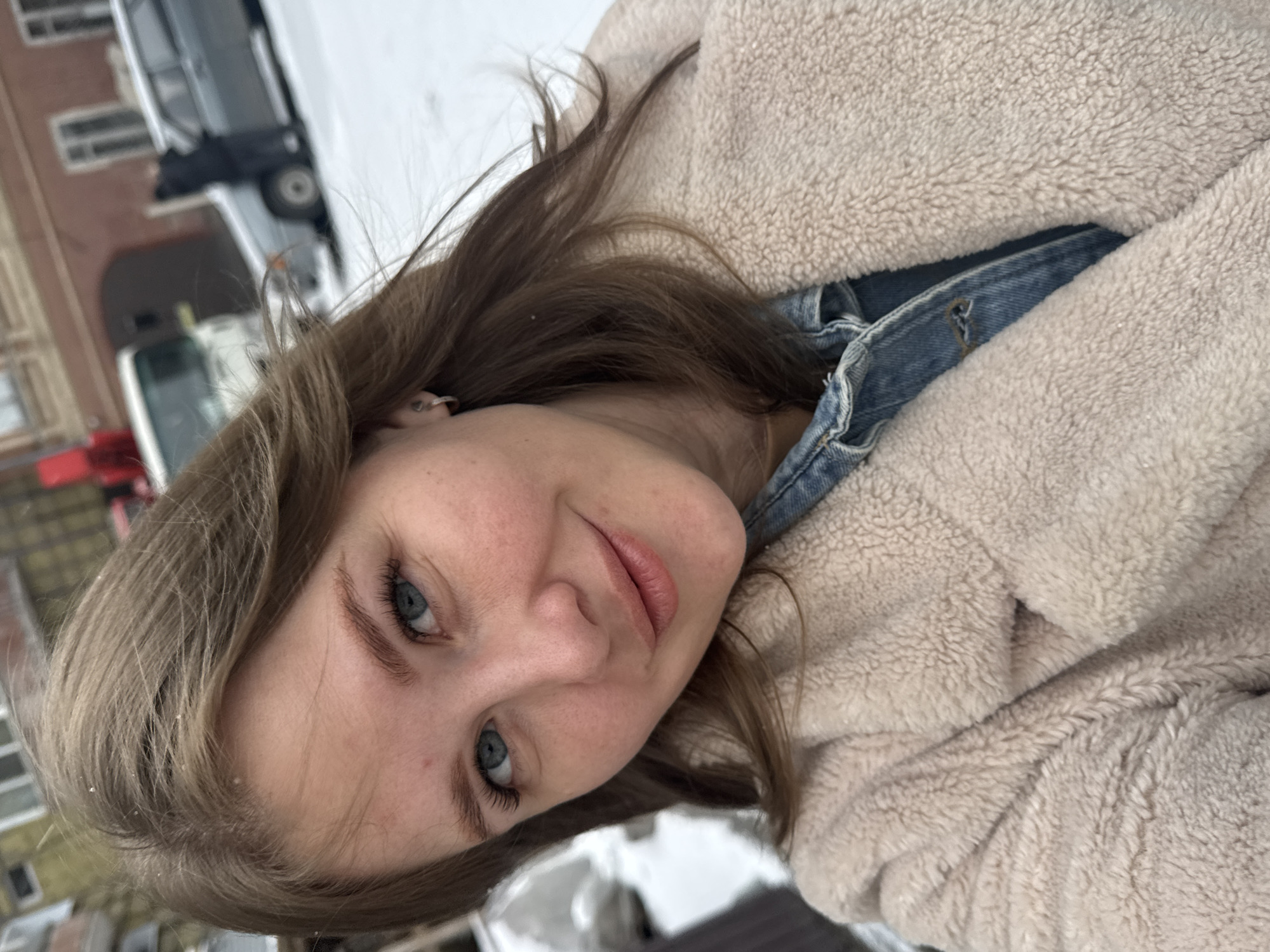 A young woman with blonde/brown hair and blue eyes poses outdoors in winter. She is wearing a beige, fleece-like jacket over a blue denim shirt or jacket. Snow is visible on her hair and in the background. The blurry background features a building with windows and parked vehicles, suggesting an urban location. Based on the information that the location is Perm, Russia, the building's architecture could potentially be of Russian style, though this is difficult to confirm due to the blur.
