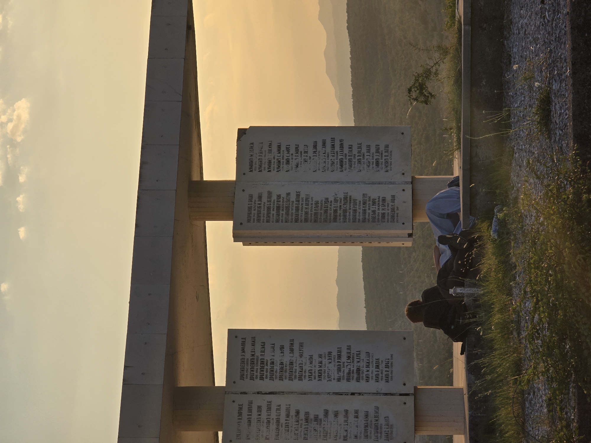 The image shows a memorial with inscribed names and dates, likely in Podgorica, Montenegro. The memorial is made of concrete and features several large vertical slabs with rows of text. Two individuals are seen resting in front of the memorial, facing away from the camera, with one person lying down and the other sitting up. A water bottle is visible next to them. The background consists of rolling hills and a hazy sky, indicating a natural outdoor setting, possibly at sunset due to the warm lighting. The overall scene suggests a place of historical significance and quiet contemplation.