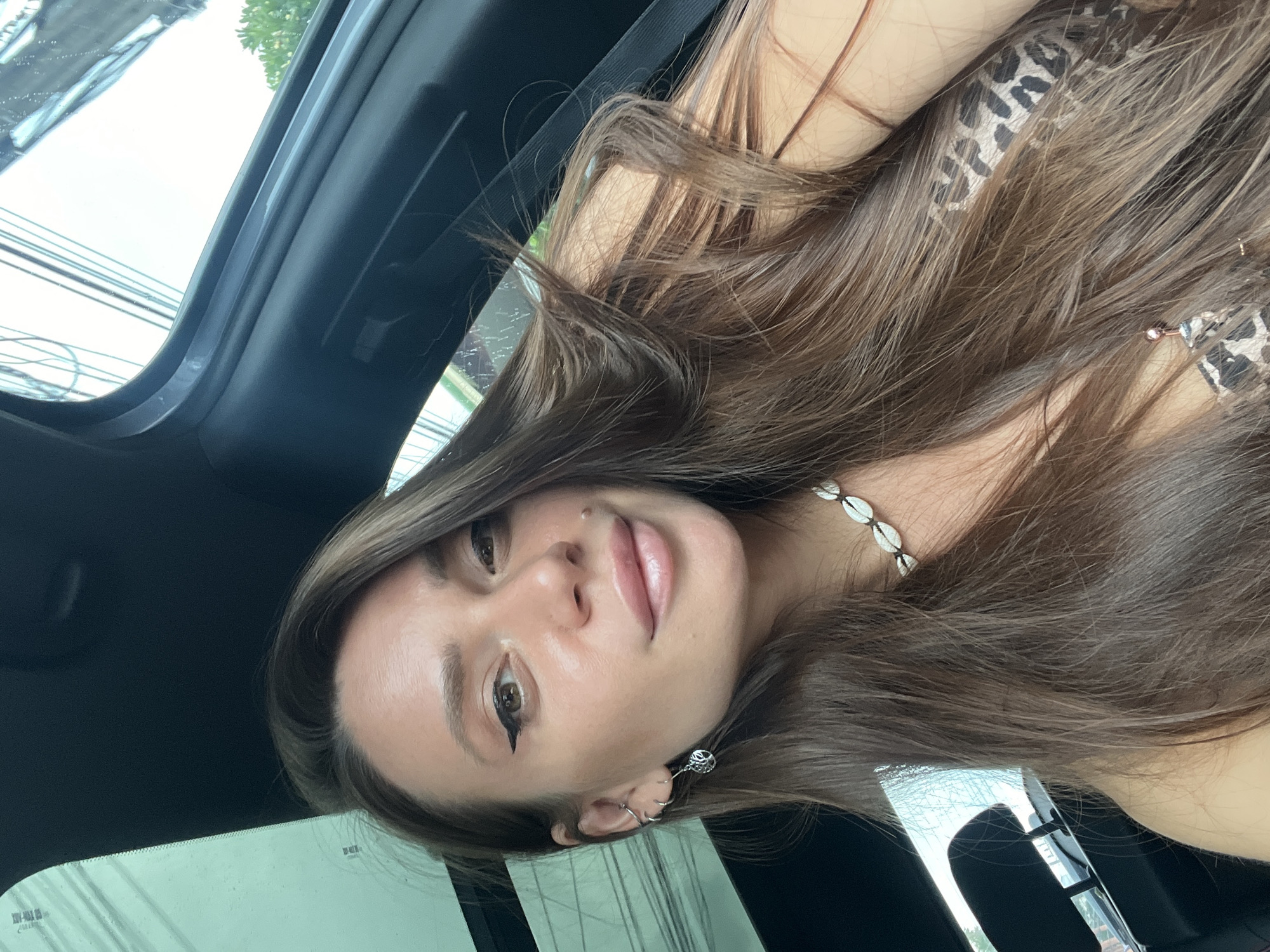 The image is a self-portrait of a young woman inside a vehicle, likely a car. The photo is rotated 90 degrees counter-clockwise.
The woman is the primary subject, with long, straight, dark brown hair falling around her shoulders and chest. She has brown eyes and is wearing winged eyeliner. A small piercing or mole is visible above her upper lip, and she has a gentle, closed-mouth smile directed towards the camera. She is wearing a top with a leopard print pattern, visible on her right shoulder. Around her neck is a necklace made of white cowrie shells. Her left ear features multiple piercings, including a prominent spiral-shaped earring in the lobe, a small silver hoop on the helix, and another ring.
The setting is the interior of a car, with dark-colored roof lining and structural elements visible. Through the car's windows, a bright, overcast sky can be seen, along with distant green trees and a network of overhead power lines, suggesting a daytime, possibly urban or suburban, outdoor environment. The lighting indicates it is daytime. On one of the car windows, faint, unreadable text or markings are visible, possibly manufacturer's labels.