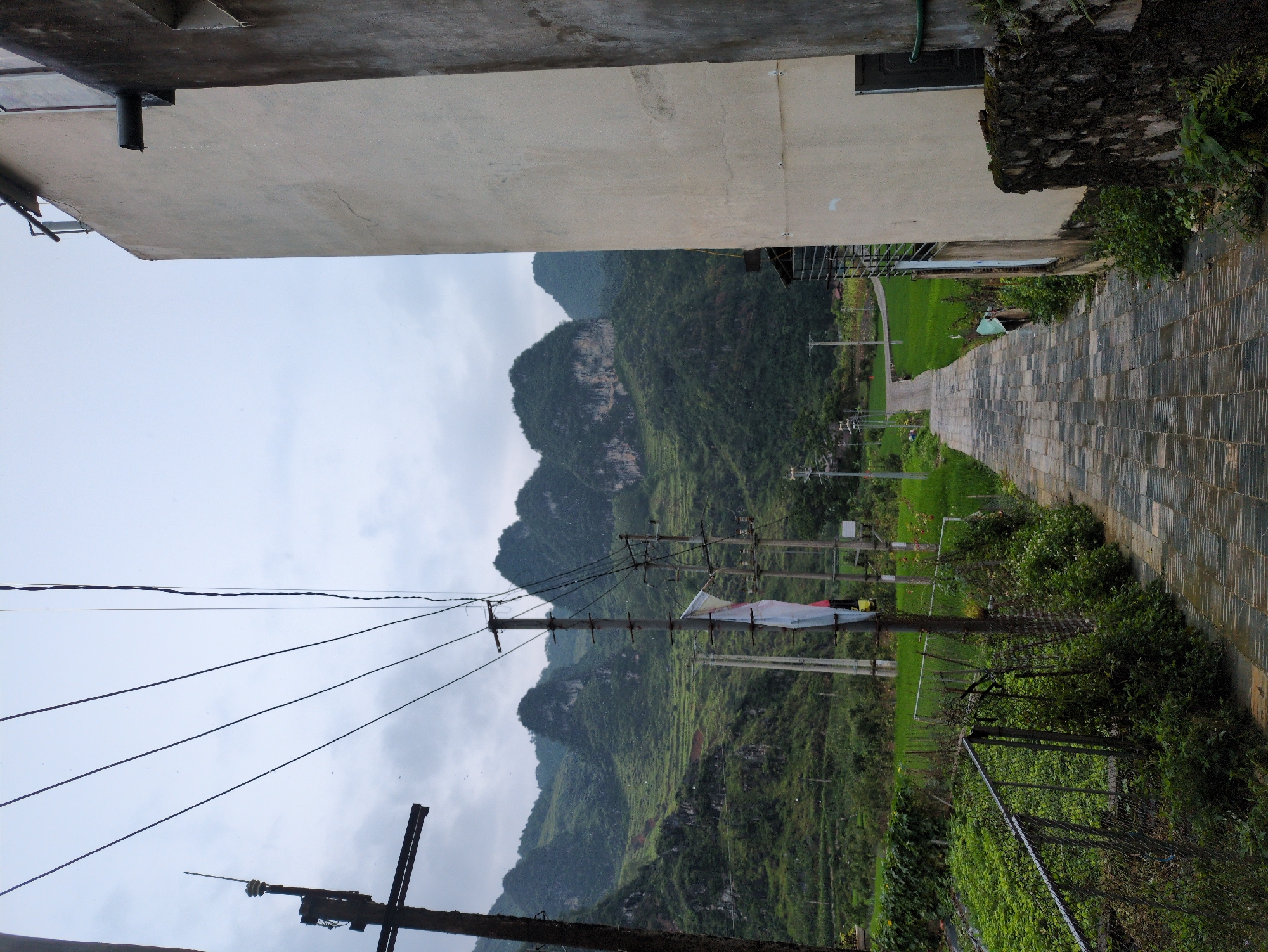 This is a vertical, eye-level, medium shot of a wet, paved pathway leading through a rural landscape in Đồng Văn Commune, Vietnam, under an overcast sky. The pathway, composed of rectangular, dark gray tiles with a textured surface, curves gently upwards and to the left, disappearing into the distance. On the right side of the pathway, a tall, beige building with a rough stucco finish dominates the foreground, with a rough, moss-covered stone wall at its base. A dark, cylindrical downspout extends from the building, with a few water droplets visible. To the left of the pathway, a chain-link fence encloses a lush green field of crops. Beyond the field, terraced hillsides covered in vibrant green vegetation rise steeply, dotted with rocky outcrops and shrouded in mist. A series of utility poles, strung with numerous electrical wires, are visible along the pathway and in the background. One pole in the mid-ground has a white and red flag attached to it. The overall atmosphere is damp and serene, suggesting recent rain. No people are visible in the frame.