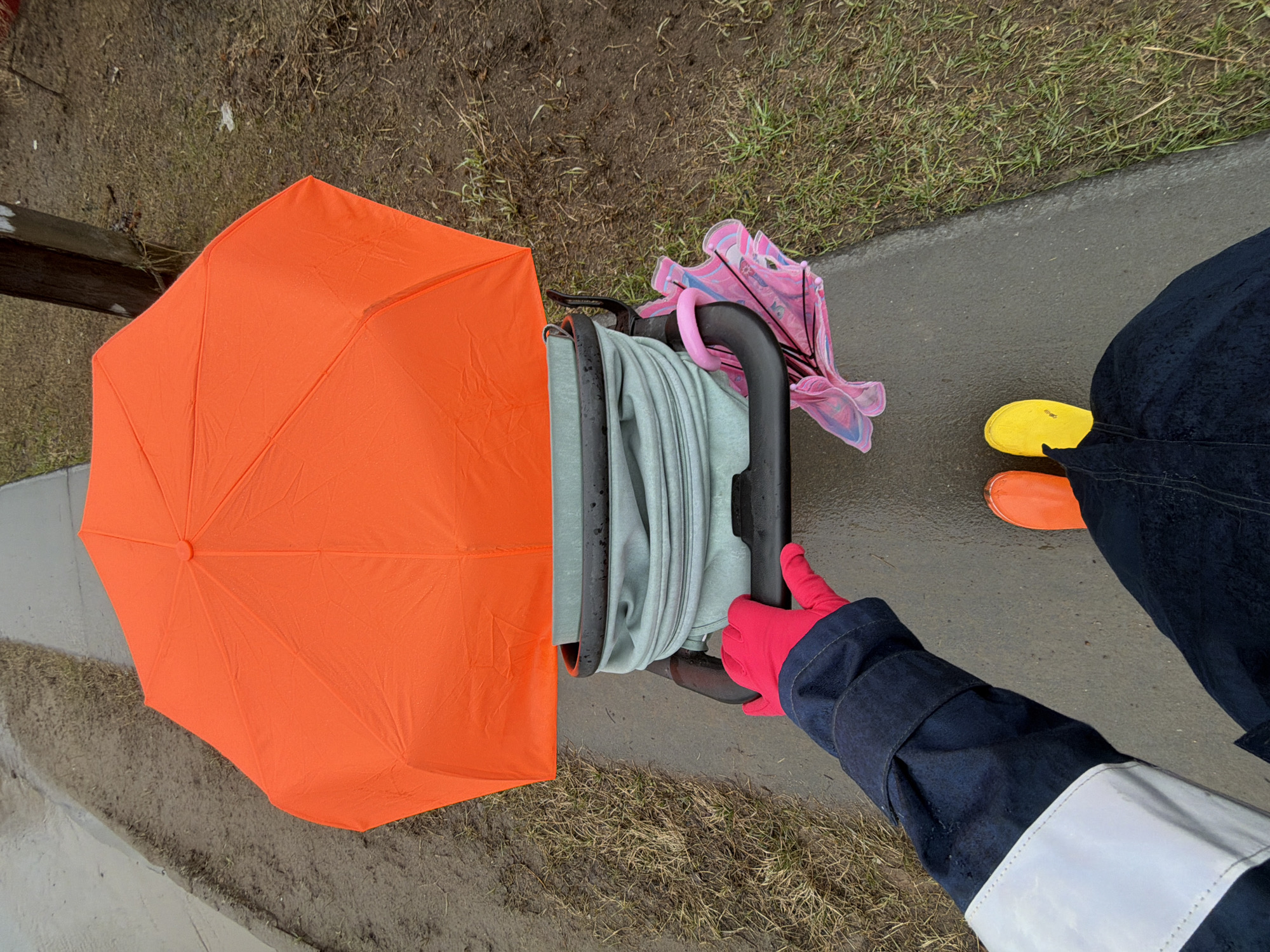 Here is a forensic description of the image:
The image presents an overhead perspective of an outdoor scene, likely taken in a wet or damp environment. Predominantly, the setting seems to be a paved walkway or sidewalk, indicated by a concrete surface. Adjacent to this pathway is an area of soil and grass. The scene includes several distinct objects and a person's arm.
An orange umbrella is prominently displayed, taking up a significant portion of the left side of the frame. To the right of the umbrella, a baby stroller or similar wheeled conveyance is visible, containing folded light blue material. A collapsed pink umbrella is attached to the stroller's handle.
A person is depicted by their arm, which is clad in a dark blue jacket with a white reflective stripe on the cuff. The hand is covered by a bright pink glove and is holding onto the stroller.
Part of the person's lower body is also visible on the right side of the frame. They are wearing dark blue pants, and their feet are clad in mismatched orange and yellow slip-on shoes.
Based solely on the image content, the exact location within the city of Chaikovsky, Russia, is not identifiable, and the scene is generalized, with no specific landmarks or signage to aid in precise geographical positioning.