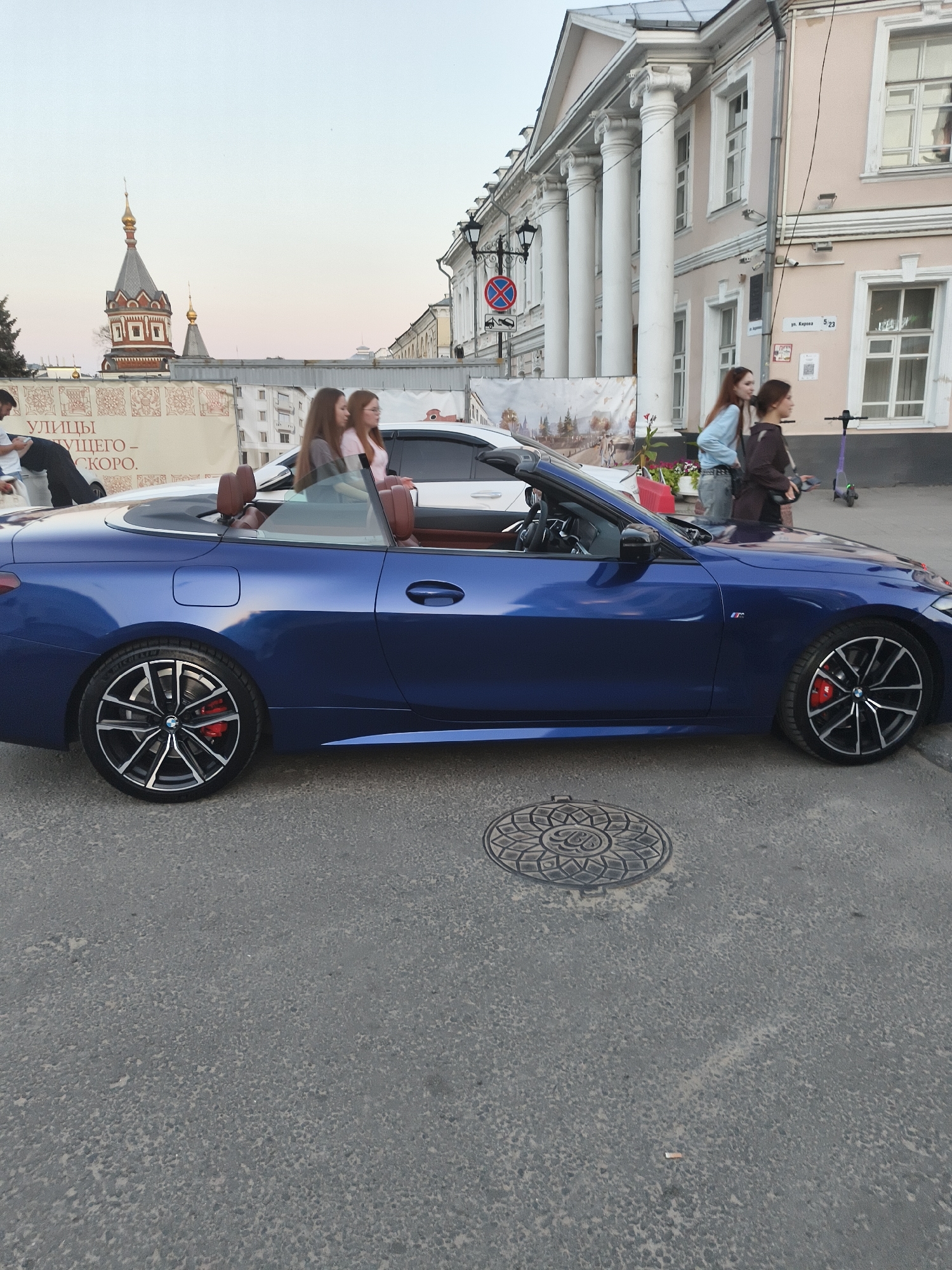 The image depicts a street scene in Yaroslavl, Russia, with a blue convertible car prominently featured in the foreground. The car is a BMW, identifiable by the M badge on its side. Inside the car, three young women are seated, appearing to be passengers. The car is parked on an asphalt road, with a manhole cover visible on the right side of the image. In the background, a light pink building with classical columns stands alongside the street. To the left, a tall, ornate building with a golden dome, likely a church or cathedral, is visible. Several people are walking on the sidewalk, and a purple electric scooter is parked near the building. A banner with Russian text and decorative patterns is also present on the left side of the image. The sky is a soft, pale blue, suggesting either early morning or late evening.