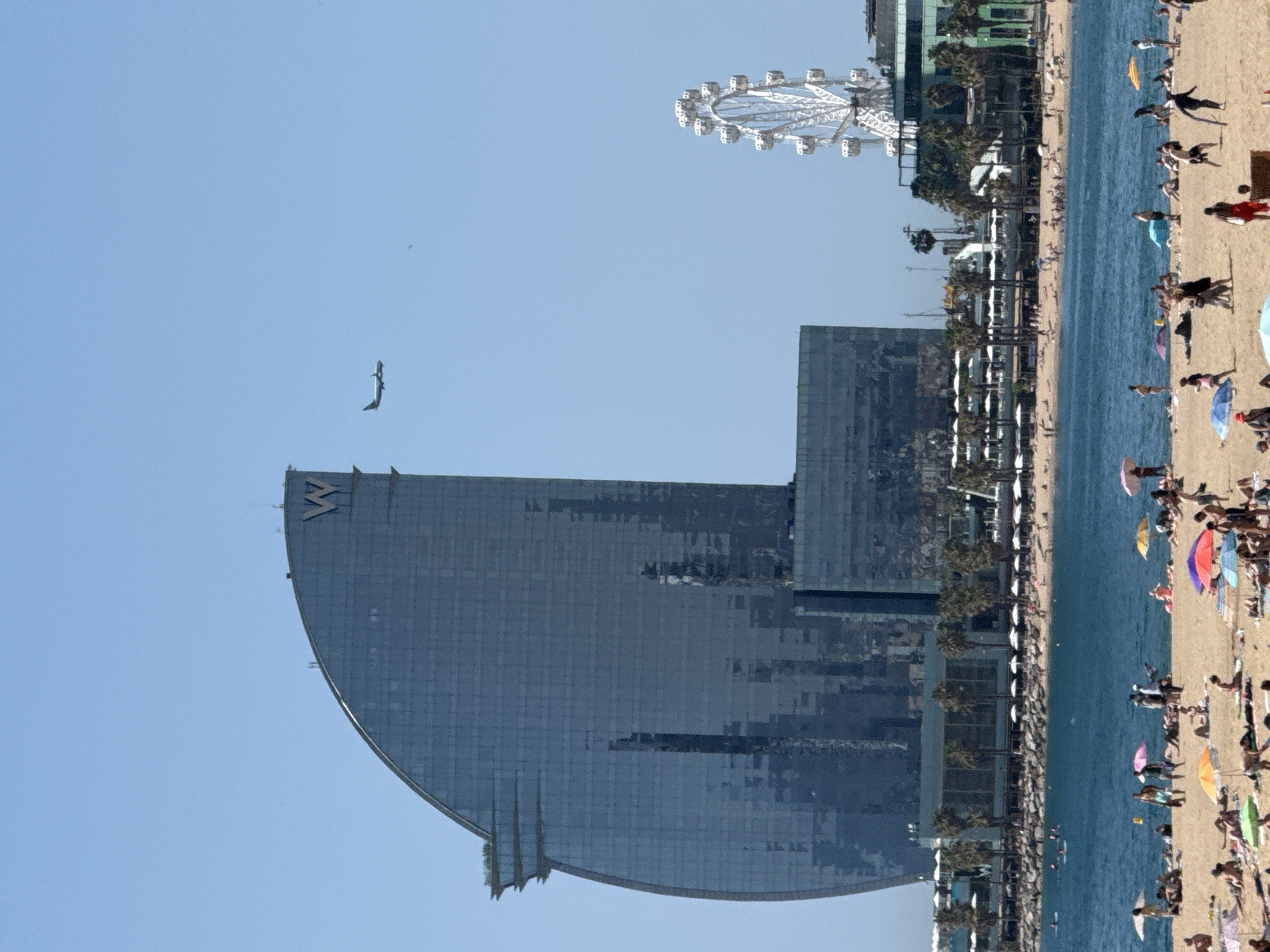 The image depicts Barceloneta Beach in Barcelona, Spain, on a clear, sunny day. In the background, the distinctive sail-shaped W Hotel dominates the skyline, with its prominent "W" logo visible at the top. To the right of the hotel, a large Ferris wheel, likely part of the adjacent Port Olímpic, is visible. The beach itself is populated with numerous people sunbathing, swimming, and relaxing under various colorful umbrellas. The blue Mediterranean Sea stretches out before the beach, dotted with small boats and people enjoying the water. Palm trees line the edge of the beach, providing some shade, and a rocky embankment separates the sand from the buildings and amenities behind it. An airplane is captured in flight in the sky above the scene.