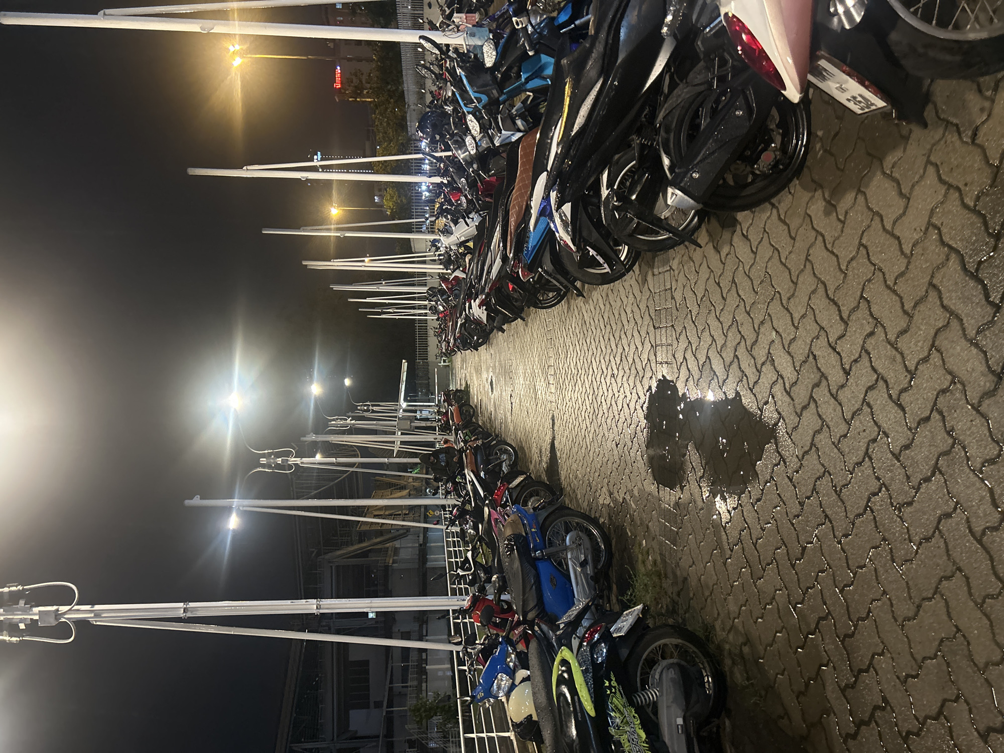 The image depicts an outdoor motorcycle parking lot at night in Tha Khlong Town Municipality, Thailand. A row of parked motorcycles is visible on a wet, patterned brick pavement. The motorcycles are predominantly of the scooter or moped type, with various colors including black, blue, red, and white. Some motorcycles have visible license plates. Tall, modern light poles with multiple fixtures line the left side of the parking area, casting illumination. In the background, there appears to be a building structure, possibly under construction or renovation, with exposed beams and scaffolding. The sky is dark, indicating nighttime. There is a single person visible in the far right of the image, wearing a dark hooded jacket and standing near a motorcycle. The overall scene is that of a functional urban space for vehicle parking at night.