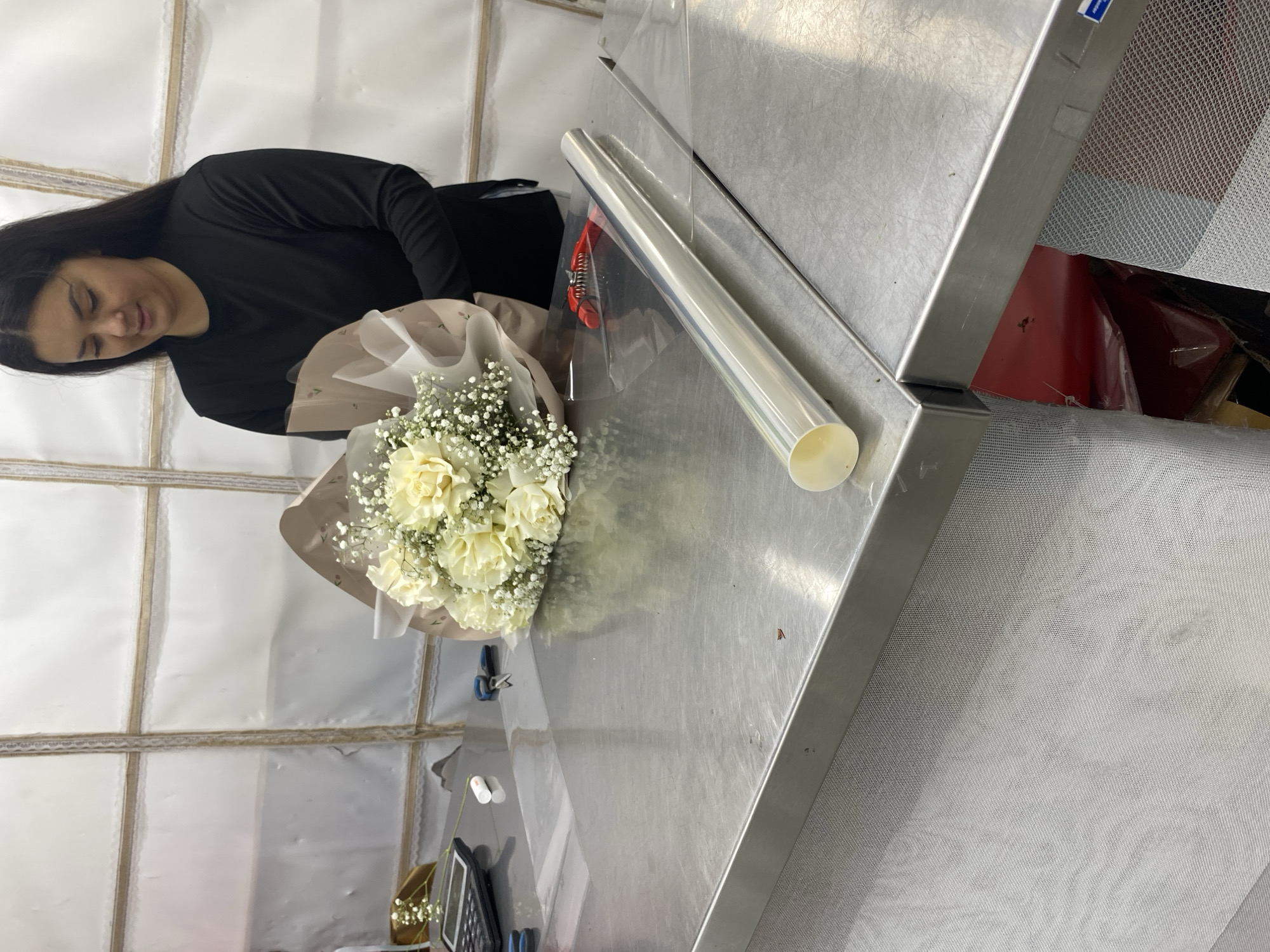 A woman with dark hair is preparing a bouquet of white flowers in an interior space. She is dressed in a black long-sleeved shirt. The bouquet is wrapped in tan-colored paper and clear cellophane. The woman appears to be working at a stainless steel counter. A roll of clear wrapping paper is on the counter near the bouquet. The background features white walls with textured accents. The location is potentially a flower shop or event-related business, but it is not possible to confirm the specific location within Yekaterinburg, Russia, based on the given image alone.