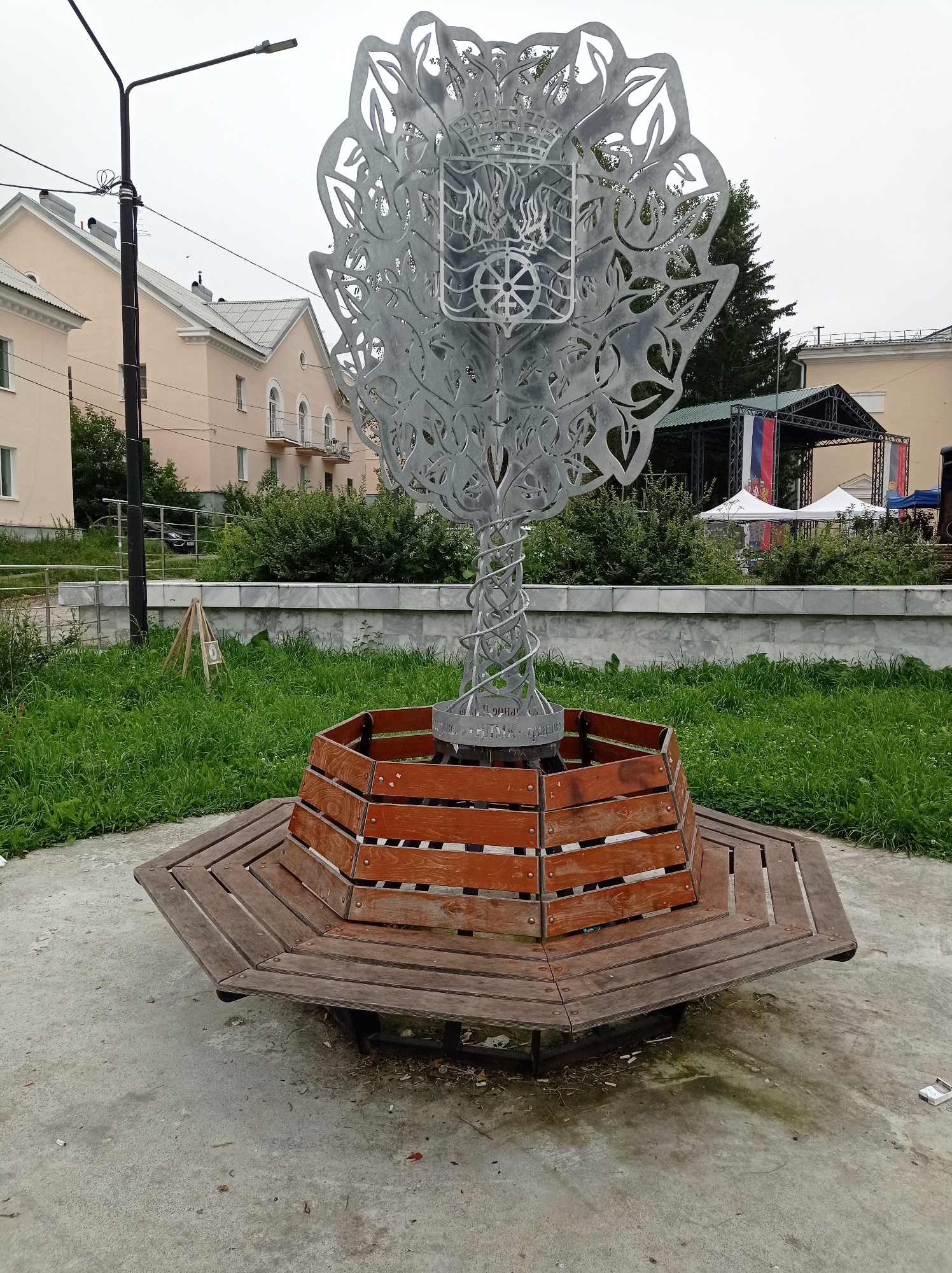 The image shows a metal sculpture resembling a tree with an intricate, ornamental design at the top, including a crest and circular elements. It is situated in a paved area with a multi-sided wooden bench surrounding its base. The bench is constructed from individual wooden planks arranged radially. Behind the sculpture and bench, there is a manicured lawn with a low marble-effect wall. Further back, buildings with light-colored facades and pitched roofs are visible, along with a street lamp. In the background, a stage with Russian flags and white tents can be seen, suggesting an event or gathering is taking place in the city of Degtyarsk, Russia. The sky is overcast.