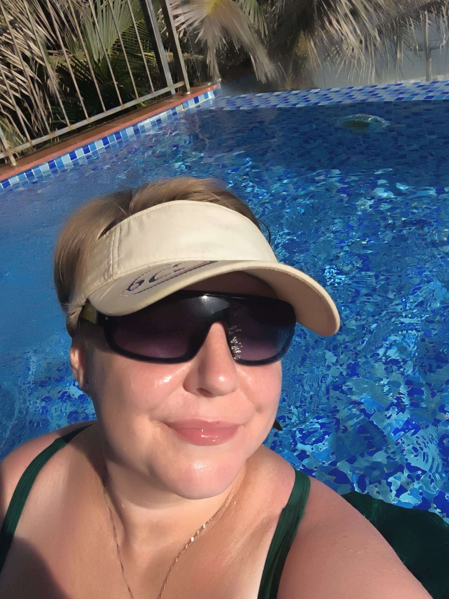A woman is taking a selfie in a swimming pool. She is wearing a green bikini top, a beige visor, and sunglasses. Her skin appears to be lightly tanned and glistening with water. The swimming pool is filled with clear blue water, and the bottom is tiled with blue and white squares. Palm trees and a fence are visible in the background, suggesting an outdoor setting, possibly in a tropical or subtropical climate. The woman's face is close to the camera, and she is smiling. The water ripples around her, indicating she is partially submerged or moving within the pool. The lighting suggests it is daytime, with bright sunlight casting reflections on the water and the woman's skin. The scene evokes a sense of leisure and relaxation.