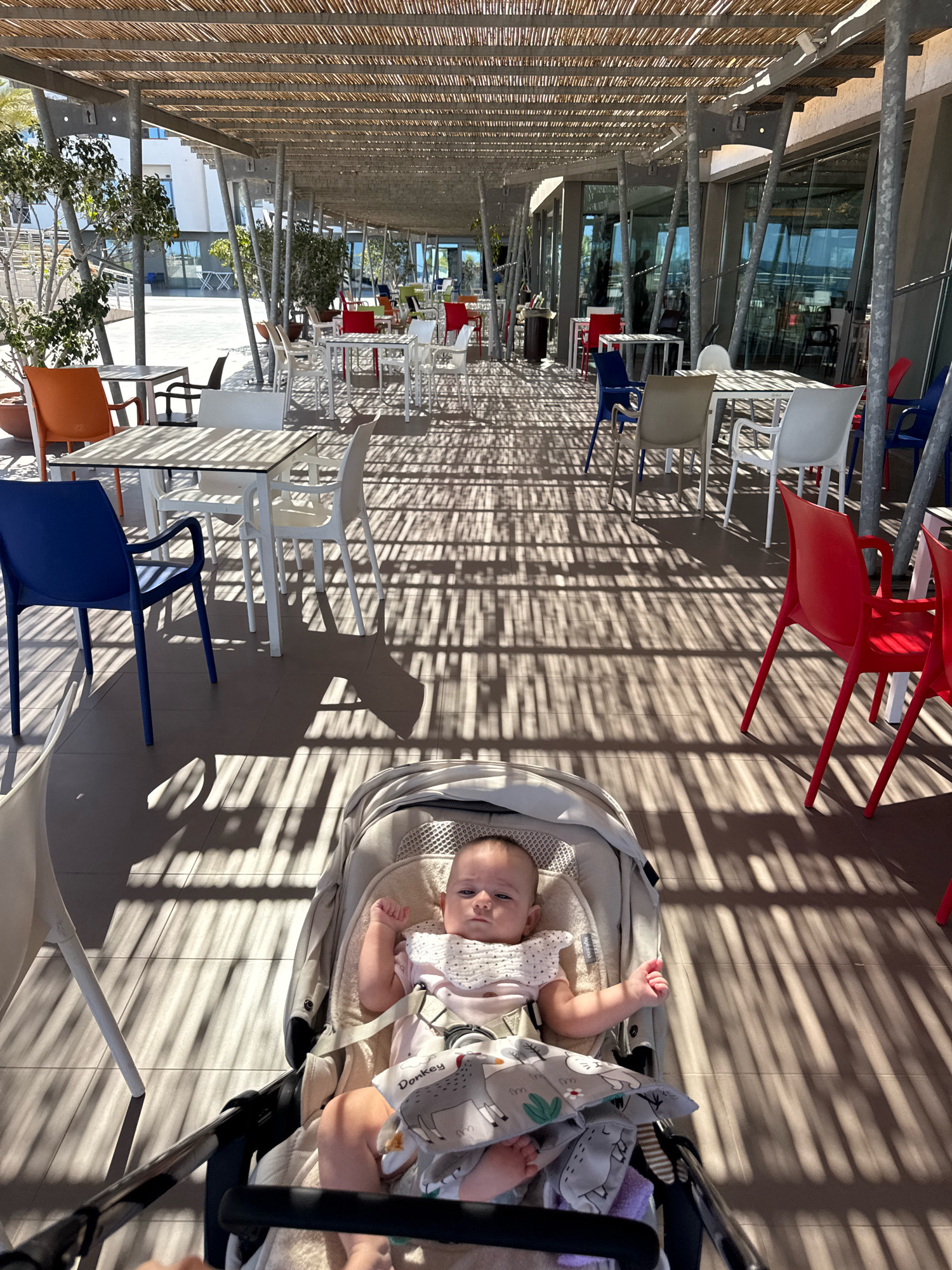 An overhead shot shows a baby in a stroller in the foreground of an outdoor dining area with tables and chairs. The baby is lying on its back and wearing a pink outfit and a bib with a pattern of animals. The stroller is light-colored and sits on a tiled floor. In the background, there are several tables and chairs, some of which are brightly colored, with a covered walkway overhead. The walkway's bamboo-like roof casts long shadows across the ground. The scene appears to be a cafe or restaurant in Isola delle Femmine, Italy, given the sunny weather and outdoor seating.