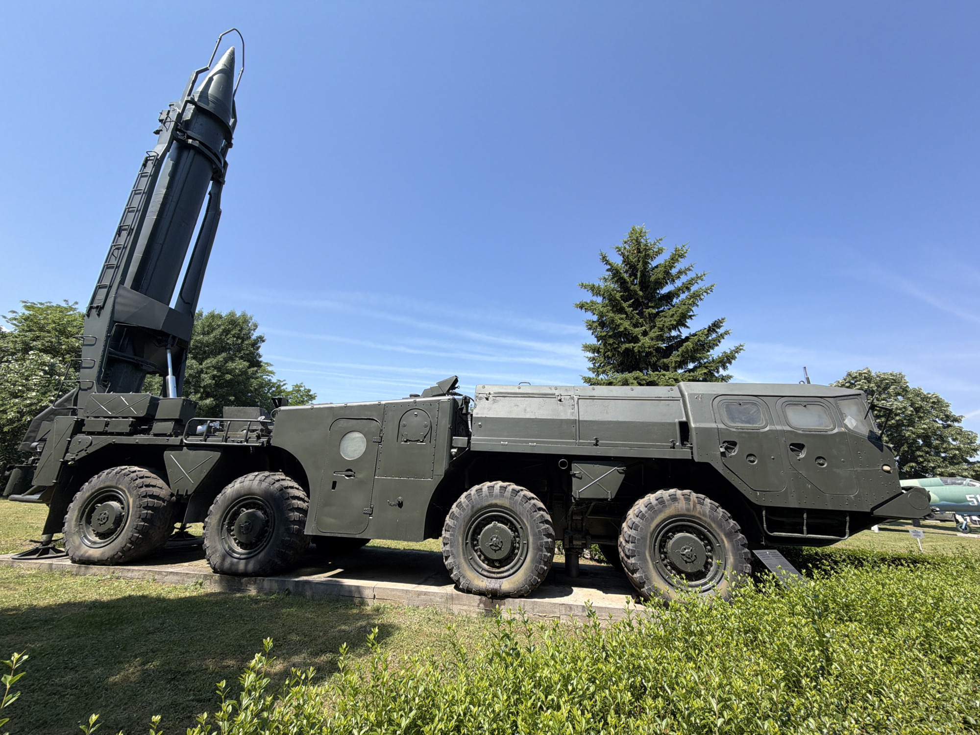 The image shows a military exhibit in the Museum of Aviation, located near Sofia, Bulgaria. In the foreground, a large, olive-drab missile launcher vehicle is displayed. The vehicle has multiple large, off-road wheels and a raised missile in a vertical position. Behind the vehicle, a green airplane with the number "51" on its tail is visible, along with other aircraft and trees. The sky is clear blue. The ground is covered with grass and some bushes in the foreground.