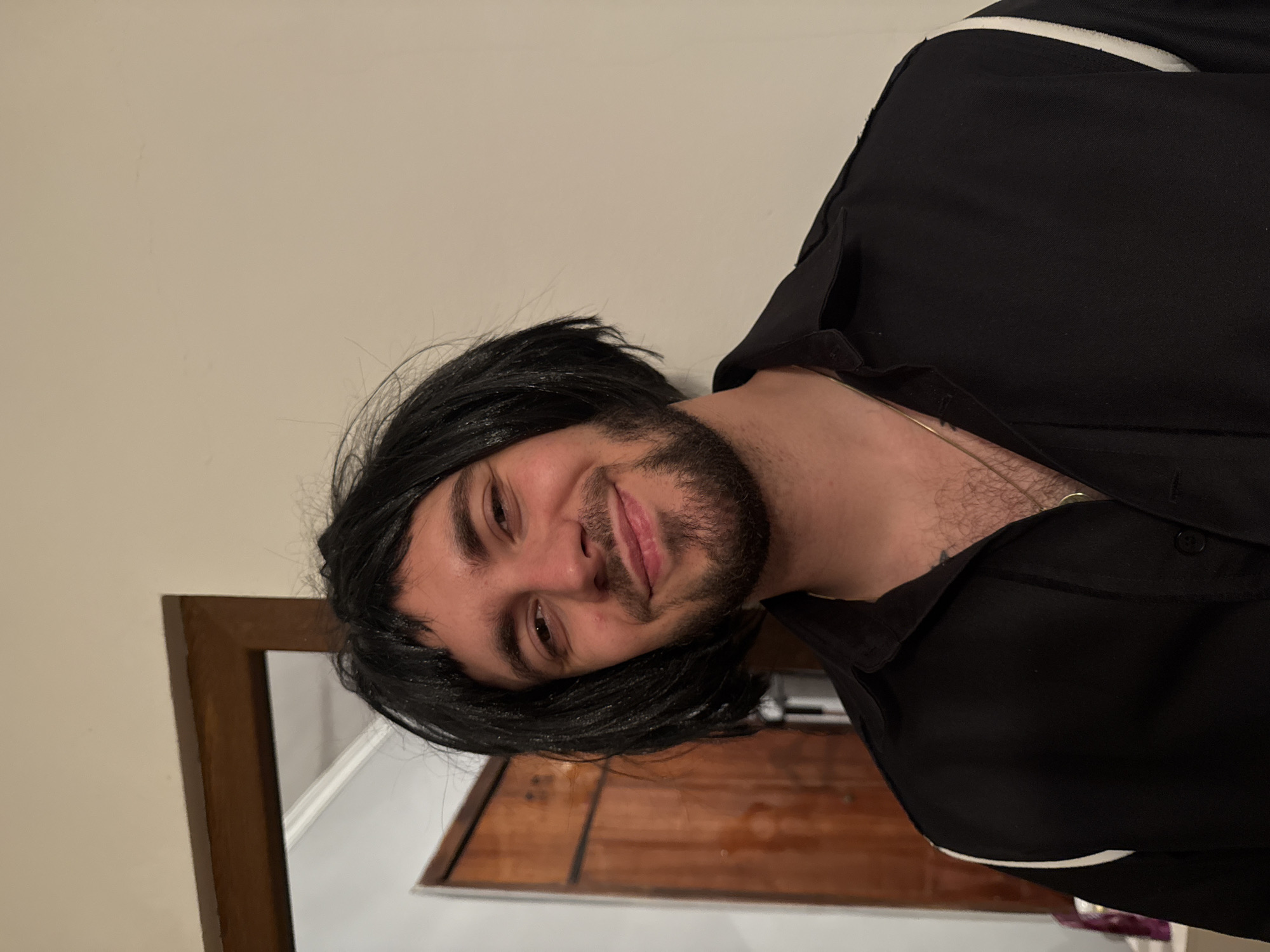 A medium shot depicts a man wearing a black wig and a black button-down shirt. The man is smiling and facing toward the viewer. The background consists of a beige wall and a wooden doorframe. It is not possible to infer the location of Yerevan, Armenia from this image.