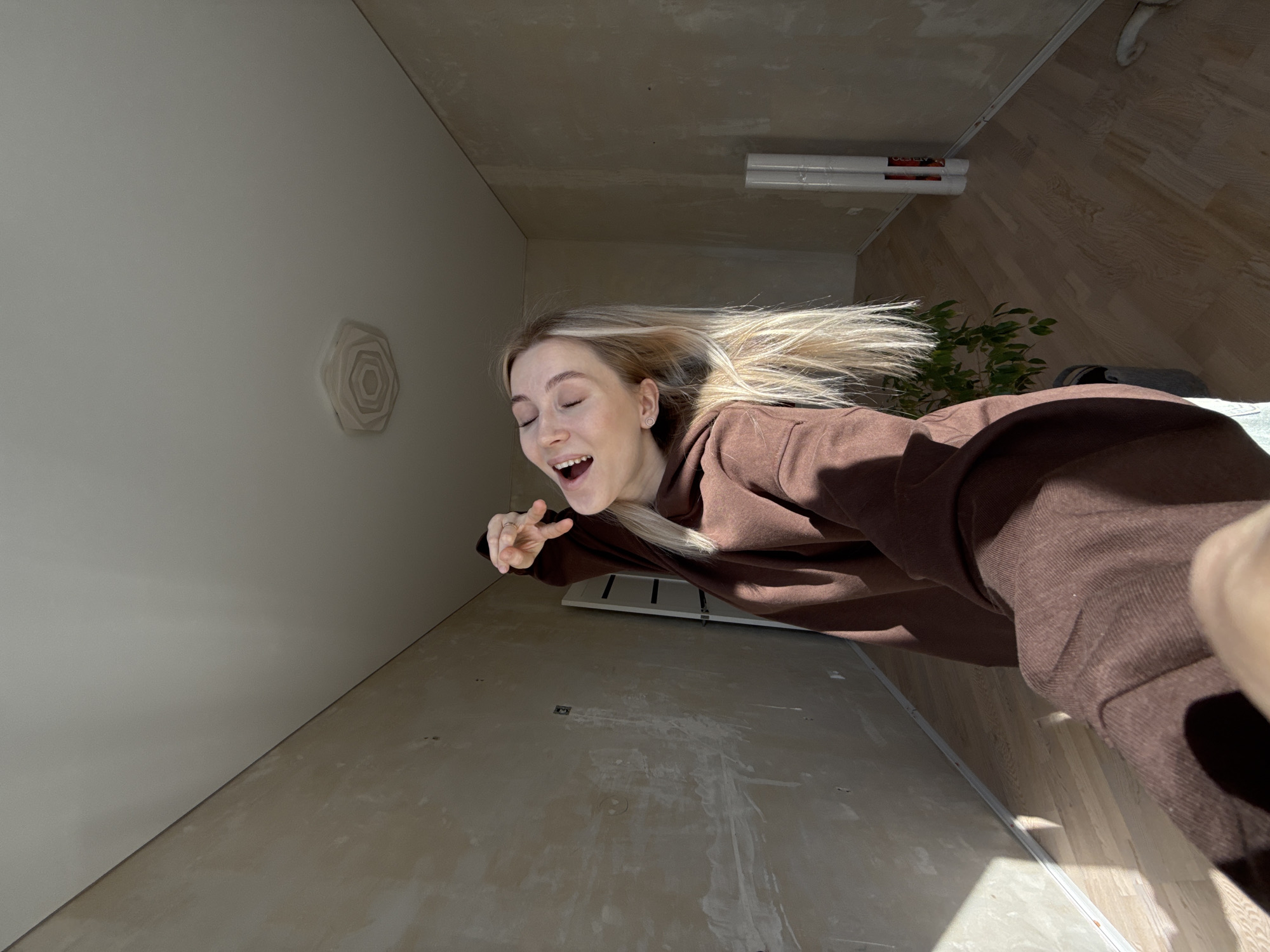 A young woman with blonde hair, wearing a brown hoodie, is angled to the right in an interior shot. The setting appears to be an apartment or home undergoing renovation. The walls are in varying states of finish, with one painted white and another with exposed plaster or cement. There's a decorative, rose-shaped item on the white wall. The floor is also unfinished. A light fixture is affixed to the ceiling. A plant and what looks like a storage heater are visible in the background. The woman's expression is one of excitement or surprise. There is insufficient information to ascertain the location in Izhevsk, Russia.