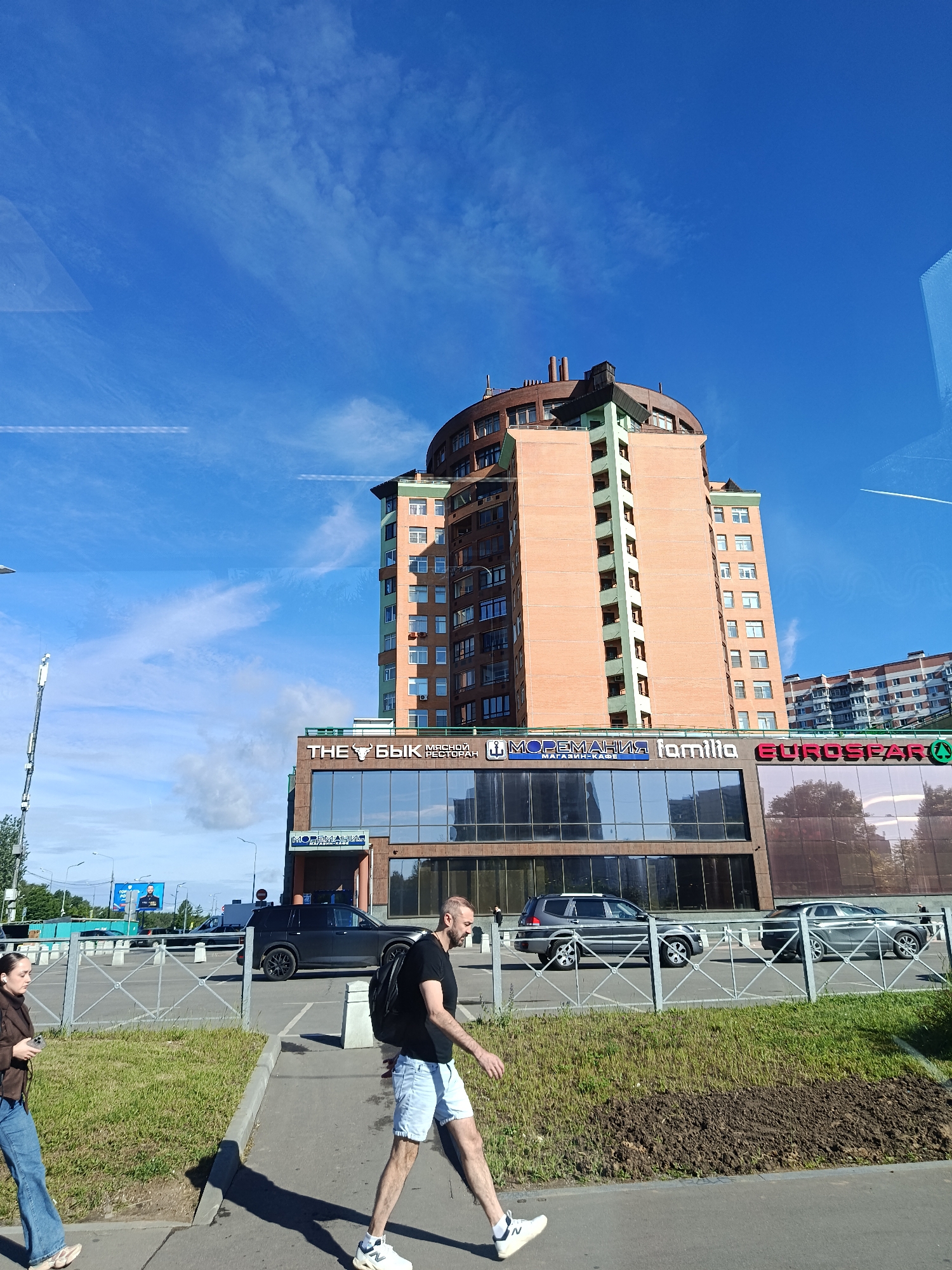 In an outdoor urban setting under a clear blue sky with scattered clouds, a multi-story brick building with a curved facade dominates the scene. The building has a mixed facade of orange brick and green accents, with multiple rows of windows and visible ventilation units. At the base of the building, large glass-fronted storefronts display signage including "THE БЫК", "МОРЕМАНІЯ", "familia", and "EUROSEAR". Two individuals are walking on a paved sidewalk in the foreground. A man with a backpack is walking towards the right, wearing a black t-shirt and light-colored shorts, and white sneakers. A woman with dark hair is walking towards the left, wearing a brown jacket and blue jeans, and holding a mobile phone. Several vehicles, including SUVs and sedans, are parked and visible behind a metal fence separating the sidewalk from a parking area. The environment includes green grass and a dirt patch.