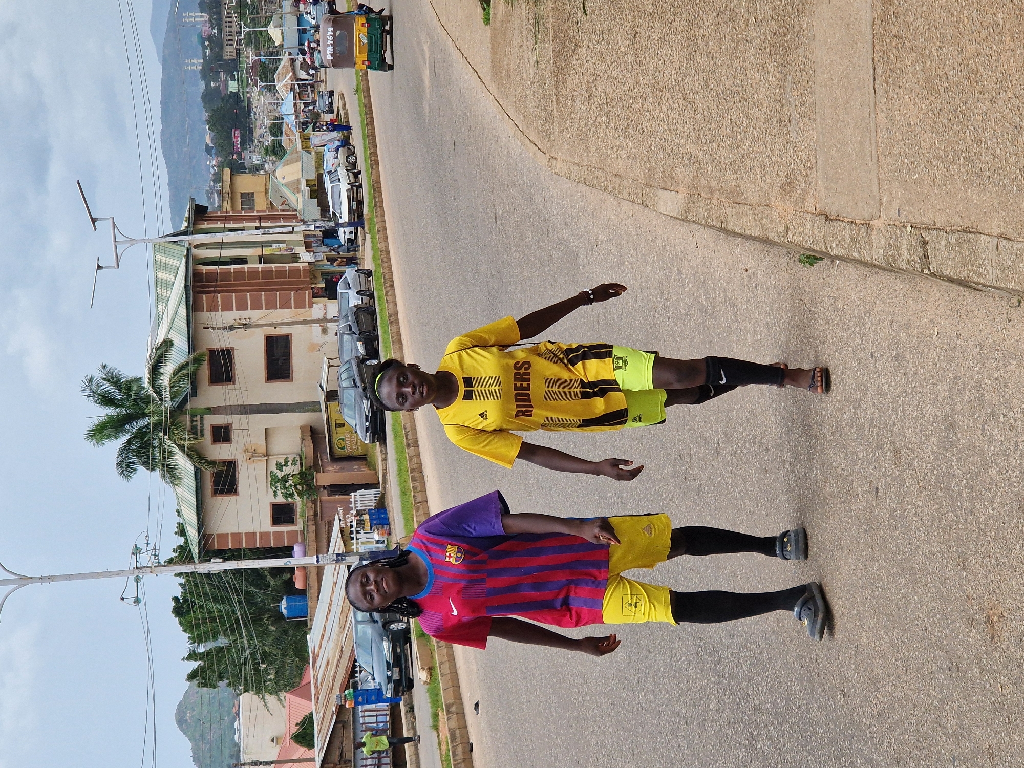 Two young women walk along a paved road in Kabong, Nigeria. The woman on the left wears a striped red, blue, and purple soccer jersey and yellow shorts with black socks and dark sandals. The woman on the right wears a yellow soccer jersey with black stripes and "RIDERS" printed in yellow, yellow shorts with black trim, and dark flip-flops. They are walking on the right side of the road, away from the camera. The road is lined with a concrete sidewalk and a grassy verge. In the background, there are buildings, trees, vehicles, and mountains. The sky is overcast.