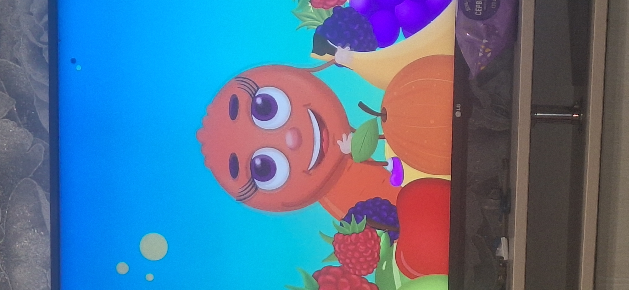 The image shows a television screen displaying a cartoon featuring a round, orange character with large purple eyes and an open smile. The character is surrounded by various cartoon fruits, including raspberries, blackberries, an apple, a pumpkin, a banana, and grapes. The background is a bright blue sky with a few light yellow circles. The television is placed on a dark gray entertainment stand, below which are two shelves. A purple bag of snacks is visible on the right side of the lower shelf. The image is taken from a low angle, suggesting the camera is positioned below the television. There is no information in the image to indicate the location in Bila Tserkva, Ukraine.