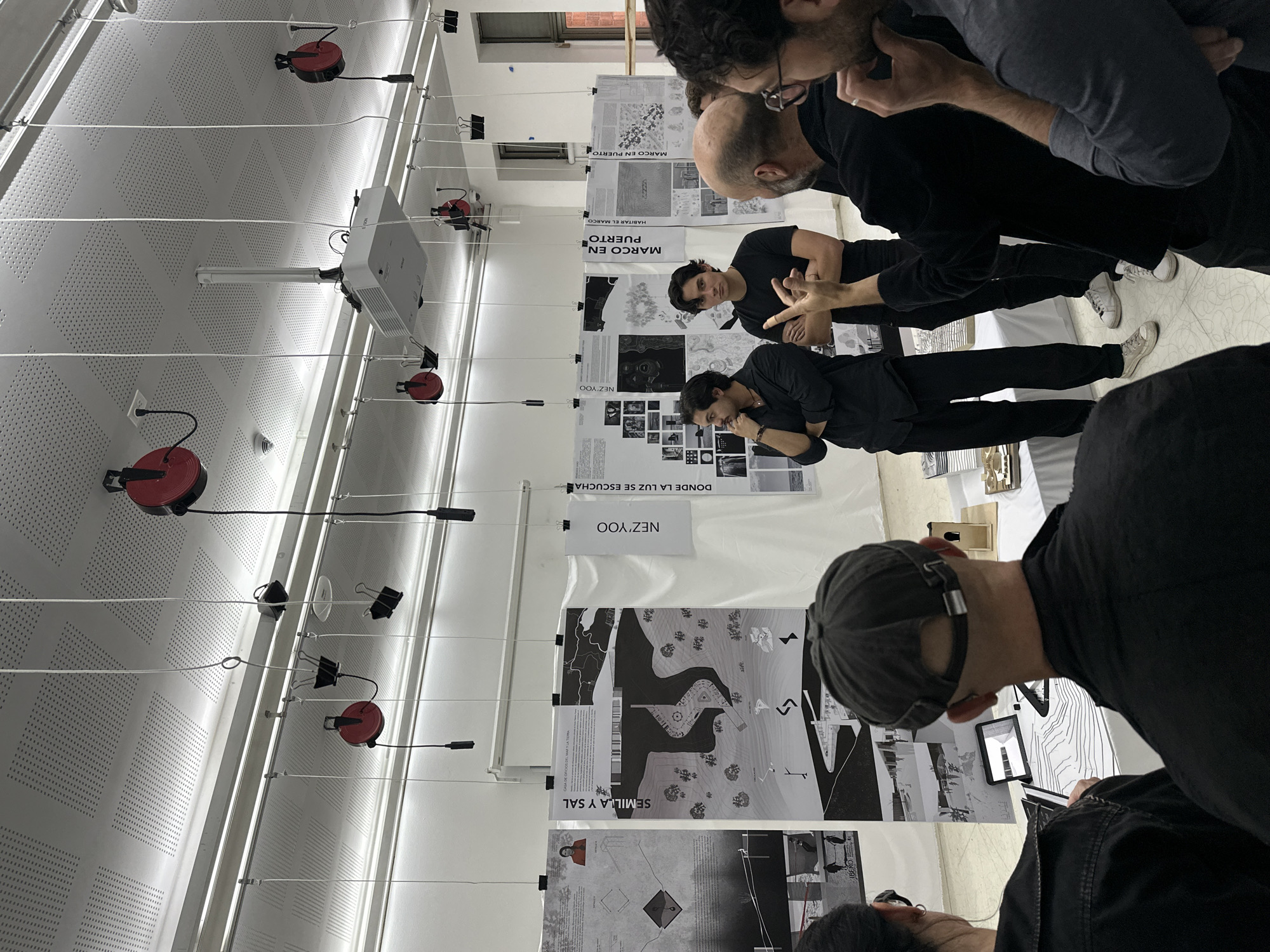 The image depicts an indoor scene in Mexico City, likely at an academic institution such as Universidad Iberoamericana, during what appears to be an architectural or design review, critique, or exhibition. Several men are gathered, intently observing and discussing large, black and white posters and potentially small models displayed on a table. Some individuals wear black clothing, and one man in the foreground has a cap and is looking down at a tablet device or a smaller display.
The setting is a brightly lit room with white walls and a white, perforated acoustic ceiling. A ceiling-mounted projector is visible, along with multiple white cables strung across the room. These cables support red retractable devices, possibly task lights or power reels, and binder clips used to hang the large format posters. The posters feature detailed architectural drawings, plans, maps, and text, indicating various design projects.
Visible text on the posters, all in Spanish, includes "IBERO", "SEMILLA Y SAL" (Seed and Salt), "CASA DE ORIC" (partially visible, possibly "Casa de Origen"), "NEZ' YOO" (appearing twice), "DONDE LA LUZ SE ESCUCHA" (Where the light is heard), "MARCO EN PUERTO" (Frame in Port / Marco in Puerto, appearing twice), and "HABITAR EL MARCO" (Inhabit the Frame). The overall impression is one of concentrated engagement and academic discussion focused on the displayed creative works.
