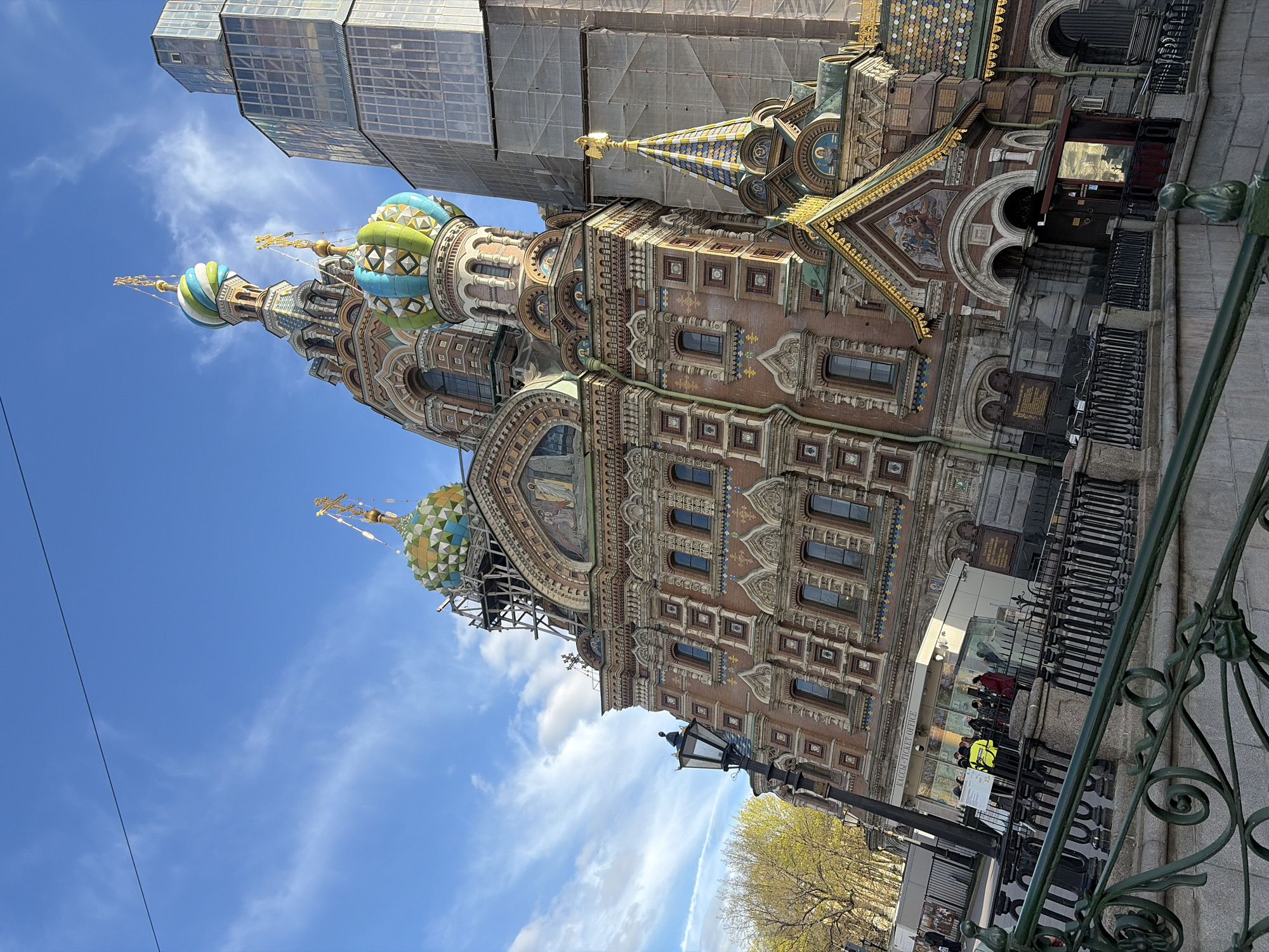 The image shows the Church of the Savior on Spilled Blood in Saint Petersburg, Russia. The ornate church, with its distinctive onion domes, is partially obscured by scaffolding and a modern building under construction. Below, a section of an ornamental iron fence, with people queuing at what appears to be a ticket office. A lamppost and some trees are also visible in the foreground, set against a blue sky with some clouds.