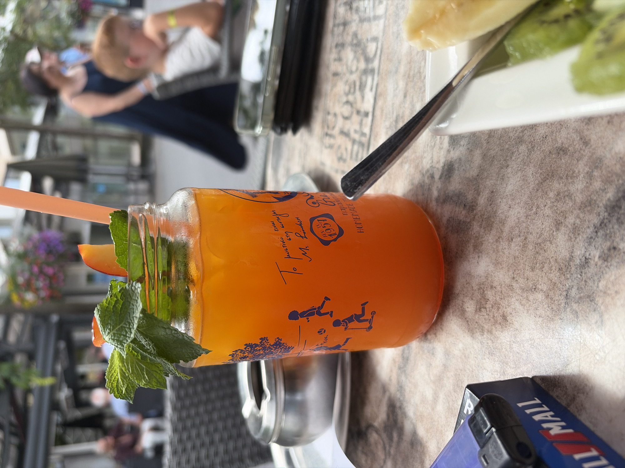In a close-up, slightly high-angle shot taken outdoors at an establishment likely in Duisburg, Germany, a glass jar filled with an orange beverage is the central focus. The jar is adorned with a blue graphic featuring children playing, one on a scooter and another kicking a ball, alongside stylized trees and text. A vibrant orange straw protrudes from the drink, topped with fresh mint leaves and a slice of fruit, possibly a peach.
To the left of the jar, a metallic ashtray sits on a table with a textured, speckled surface. In the background, the scene is blurred, but hints of an outdoor seating area are visible, with people in casual attire. A woman in a dark blue dress walks away from the camera, and a young boy with reddish hair is seated, looking down and resting his chin in his hand. Another person, out of focus, is seated behind the boy. In the far background, there are hints of greenery and structures, suggesting a public outdoor space or a cafe with an outdoor section.
In the foreground, a portion of a blue and white package, possibly a cigarette pack, is visible on the left, with the word "MALL" partially legible. To the right of the jar, a white plate holds sliced fruit, including what appears to be kiwi and a pale yellow fruit, possibly banana. A silver fork or spoon rests on the plate, with its handle extending towards the jar. The overall impression is a snapshot of a relaxed moment at an outdoor cafe, potentially during daytime given the lighting. The presence of an ashtray and smoking accessories, alongside the beverage and fruit, suggests a common leisure setting.