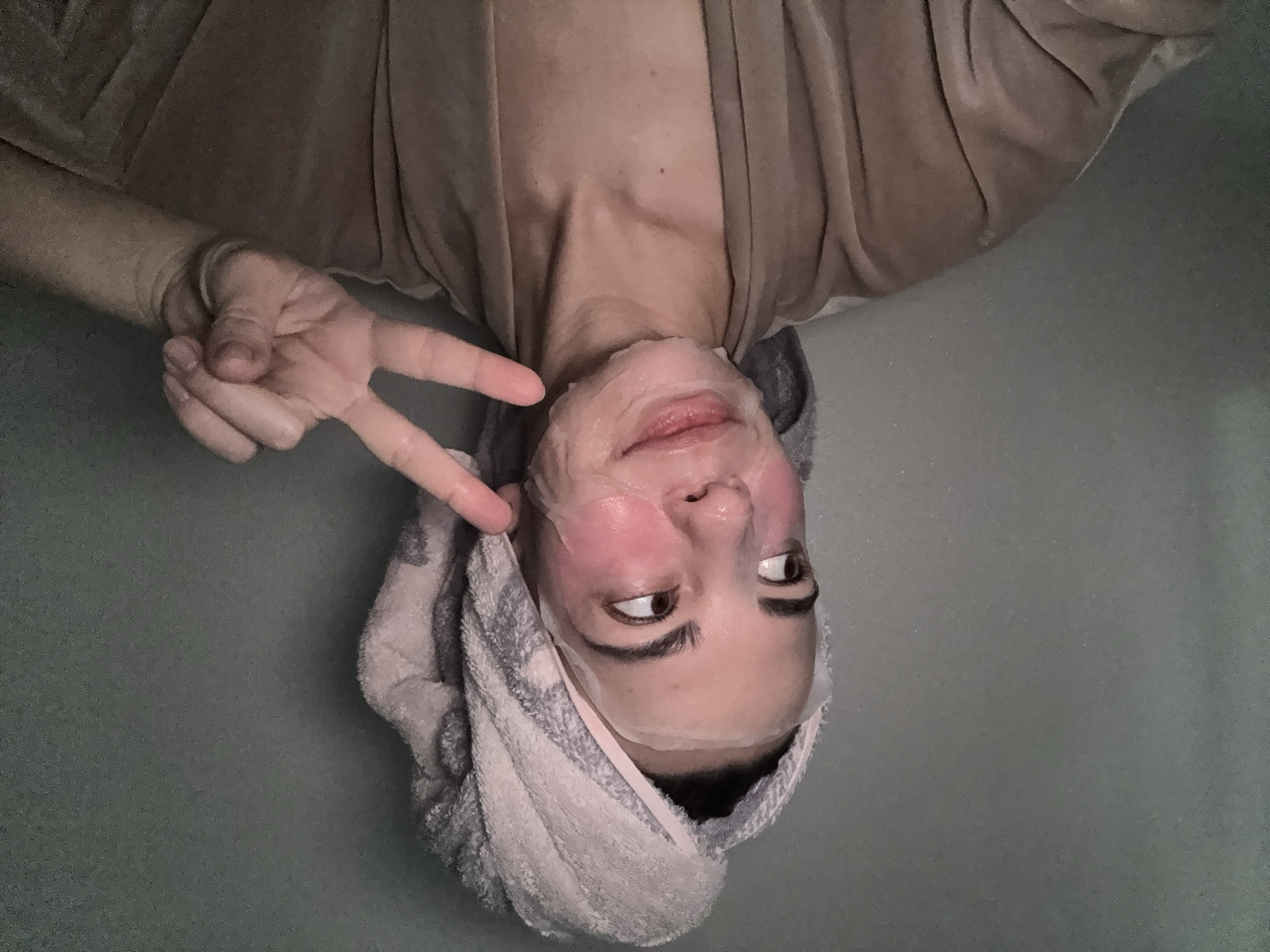 An inverted close-up shot features a person, likely a woman, lying down and looking towards the right side of the frame. Her head is positioned at the bottom of the image, wrapped in a light gray and white striped towel, suggesting a recent bath or hair washing. A translucent sheet mask covers her face, with areas on her cheeks appearing flushed or reddish. She has a slight smile on her lips and her dark eyes are directed to the side. Her left hand is raised, making a peace sign. She is wearing a soft, beige or light brown robe or garment, which is visible across her chest and arms. The background consists of a muted greenish-gray surface, possibly a bed or couch. The overall atmosphere appears relaxed and playful, with the subject engaged in a self-care activity. There is no visible text in the image.