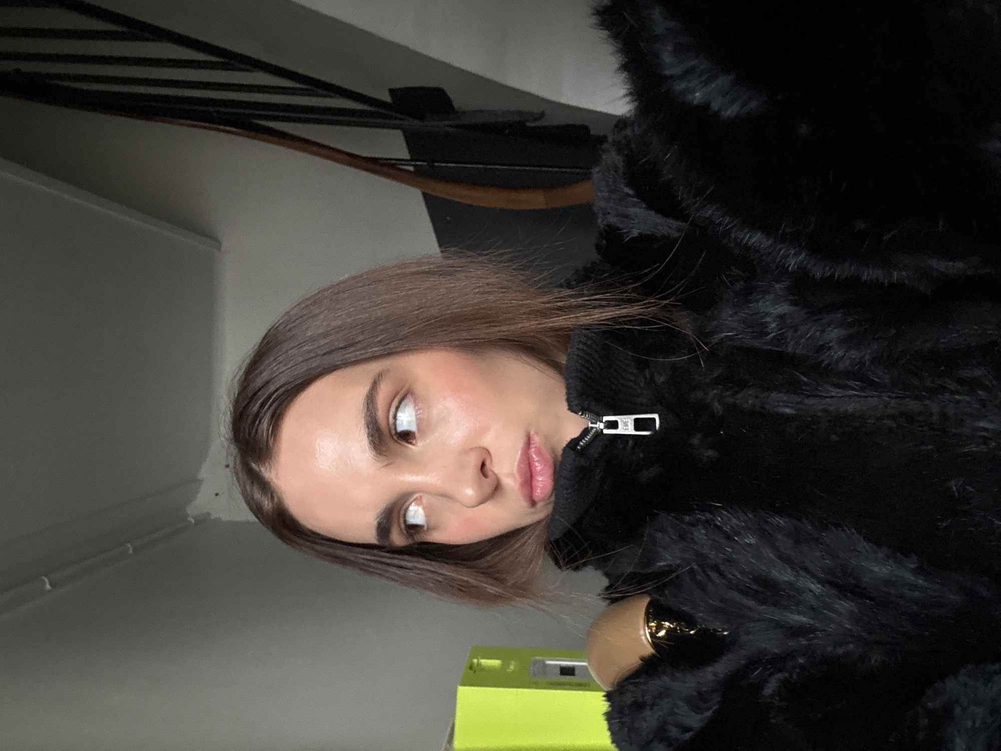 A young woman poses indoors, facing slightly left of the camera. She is wearing a black furry coat with a visible zipper pull. Her hair is straight, brown and parted to the side. Behind her is a stairway with black metal railings. The walls are painted a light grey. A bright green rectangular object is visible at the bottom of the frame. While the information required to identify the location as Paris, France is not available from the image alone, the details of the staircase architecture suggest an urban setting, which is congruent with buildings found in Paris.