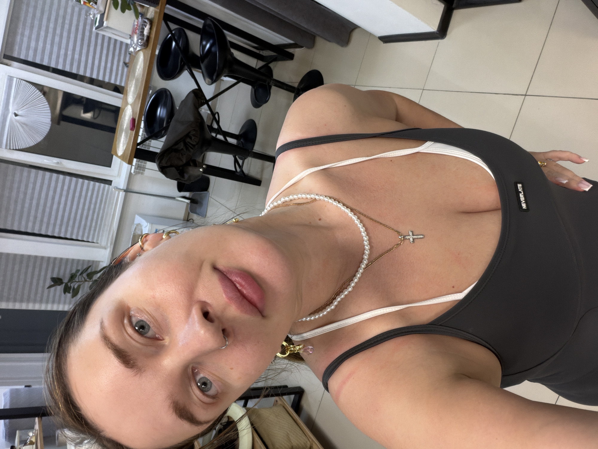A female individual is depicted in a selfie taken indoors, within what appears to be a residential kitchen or dining area in Chaikovsky, Russia. The subject is a Caucasian female with light brown hair, blue-gray eyes, and a fair complexion. She wears a silver-colored hoop nose ring on her left nostril and multiple gold-toned earrings, one of which includes a pink faceted droplet. Her attire consists of a dark gray athletic tank top, visible with the text "NEVERLATE" on the chest area, layered over a white sports bra. She wears a double-strand pearl necklace and a gold-toned chain with a small, jeweled cross pendant. Red linear marks are visible on her left and right shoulders. Her left hand is partially visible, displaying manicured nails with white tips.
The background features a light-colored tiled floor. A white-framed window with light-colored pleated blinds occupies the upper central area, with a white, fan-shaped decorative object attached to the top-left corner of the window. Below the window, a white radiator is visible. To the right, a wooden bar-style table is accompanied by three black metal and plastic bar stools. On the table are two circular, transparent placemats, a small red rectangular object, a clear plastic container holding dark-colored food items, and a small potted plant. A dark garment is draped over one of the bar stools. A dustpan and brush are visible leaning against a wall to the left of the table.