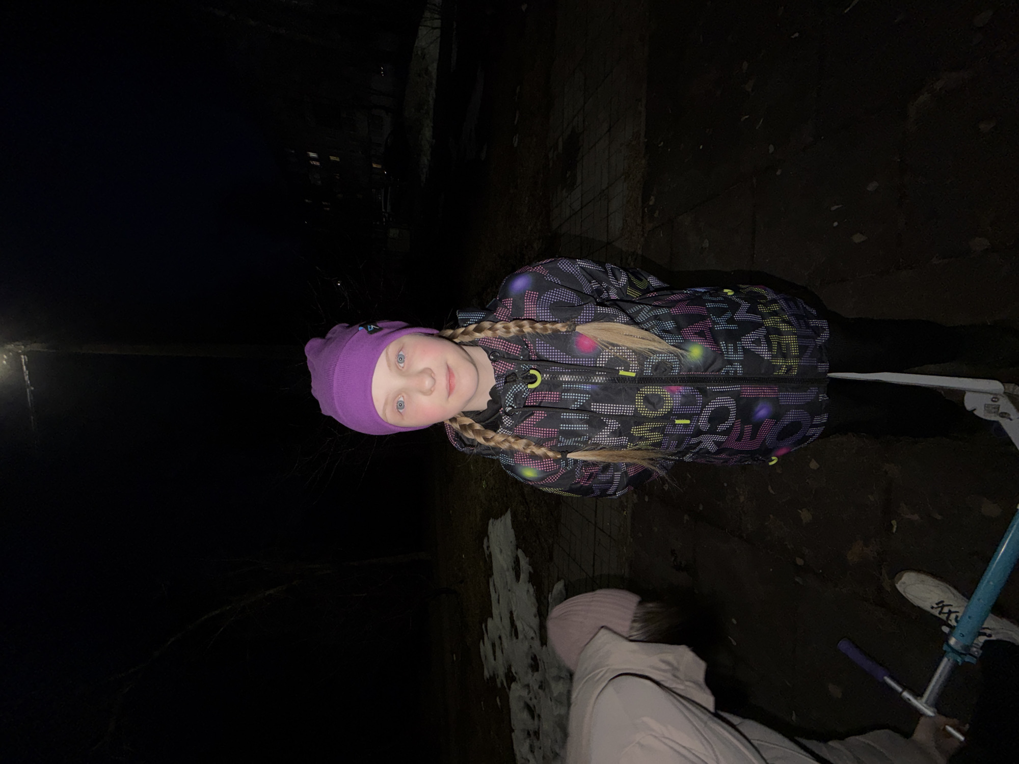 The image shows a young girl in a dark outdoor setting. The girl is wearing a purple knit hat, a patterned winter jacket, and has blonde hair styled in braids. She is looking directly at the camera. To the right of the girl, there is a scooter, and another person partially visible. The ground appears to be a mix of pavement, dirt, and patches of snow. The scene is dimly lit, with a street light visible in the distance. While I do not have enough information to definitively determine the location in the city of Chaikovsky, Russia, the presence of snow and a streetlight suggests it is an outdoor area, potentially a park or residential neighborhood.