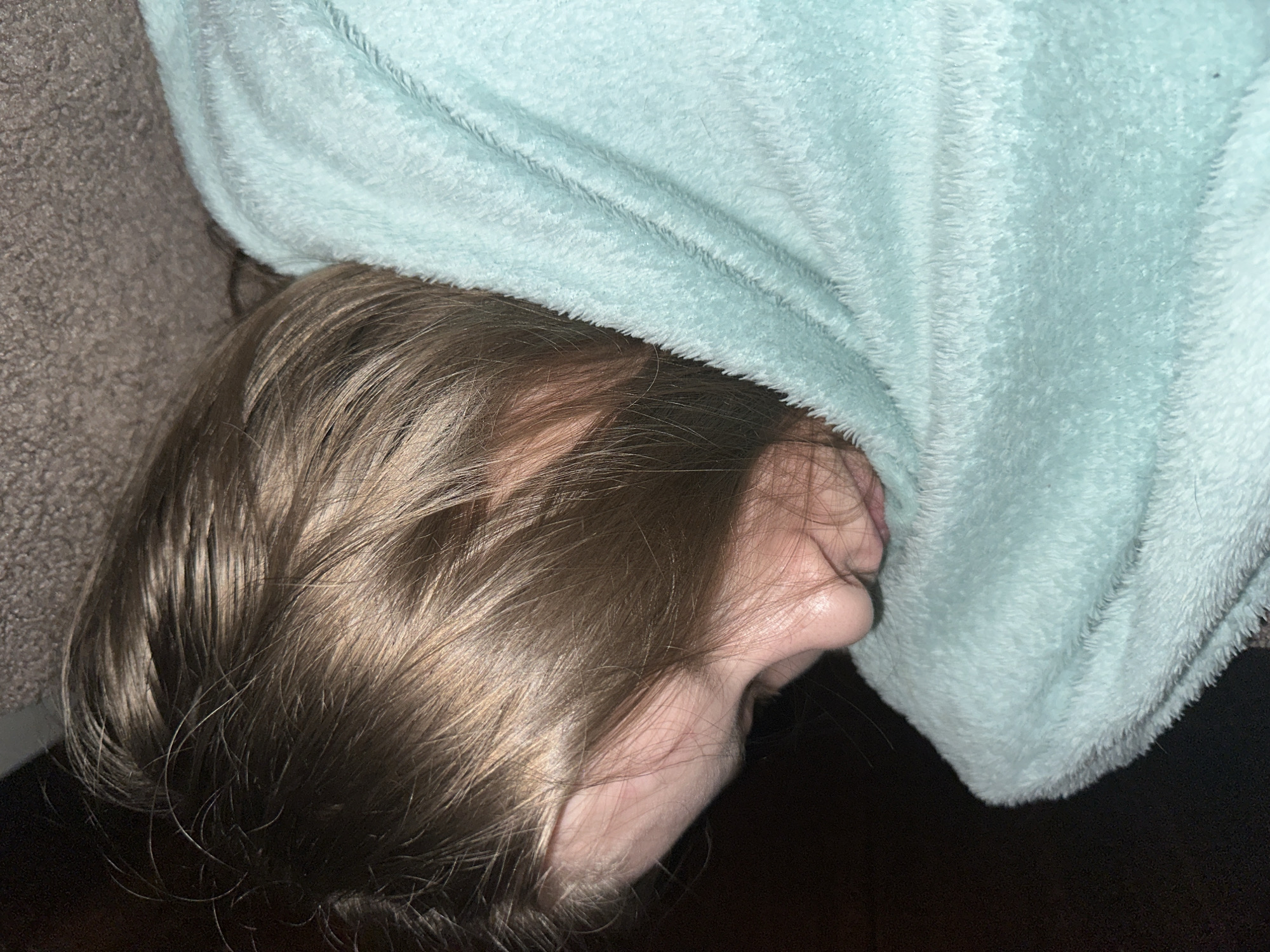 The scene depicts an individual resting indoors, located in Moscow, Russia. A person with light brown hair is visible, positioned on their right side. Their face is partially obscured by a light blue, plush, textured blanket that covers the lower portion of the face and body. The visible features include the forehead, right ear, and nose, suggesting the eyes are closed or facing away from the camera. The background consists of a dark, indistinct surface immediately behind the head, transitioning to a grey, textured surface, likely fabric upholstery, above the blanket. Lighting is subdued.