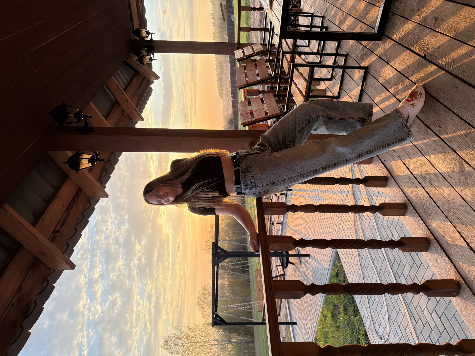 A young woman with long hair is sitting on a wooden railing in an outdoor setting. She is wearing a black crop top and light-wash jeans. Her left leg is extended onto the wooden deck, and she is wearing socks with a Christmas-themed design. The background features a sunset sky over trees. A wooden structure with lights is partially visible above her. A wooden table and chairs are also on the deck. A swing set is visible beyond the railing. The image location is possibly Kamennoye, Russia.