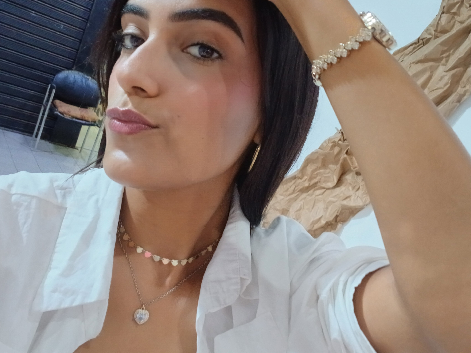 A woman with dark hair is posing for a selfie. She's wearing a white, unbuttoned collared shirt, a gold heart-shaped pendant necklace with a second, shorter necklace of small gold hearts, and a gold watch or bracelet on her raised arm. Her makeup is subtle, with pinkish lipstick and blush. The background includes a closed metal shutter, a blue chair, and crumpled brown paper. The photo appears to be taken indoors, possibly in Porlamar, Venezuela.