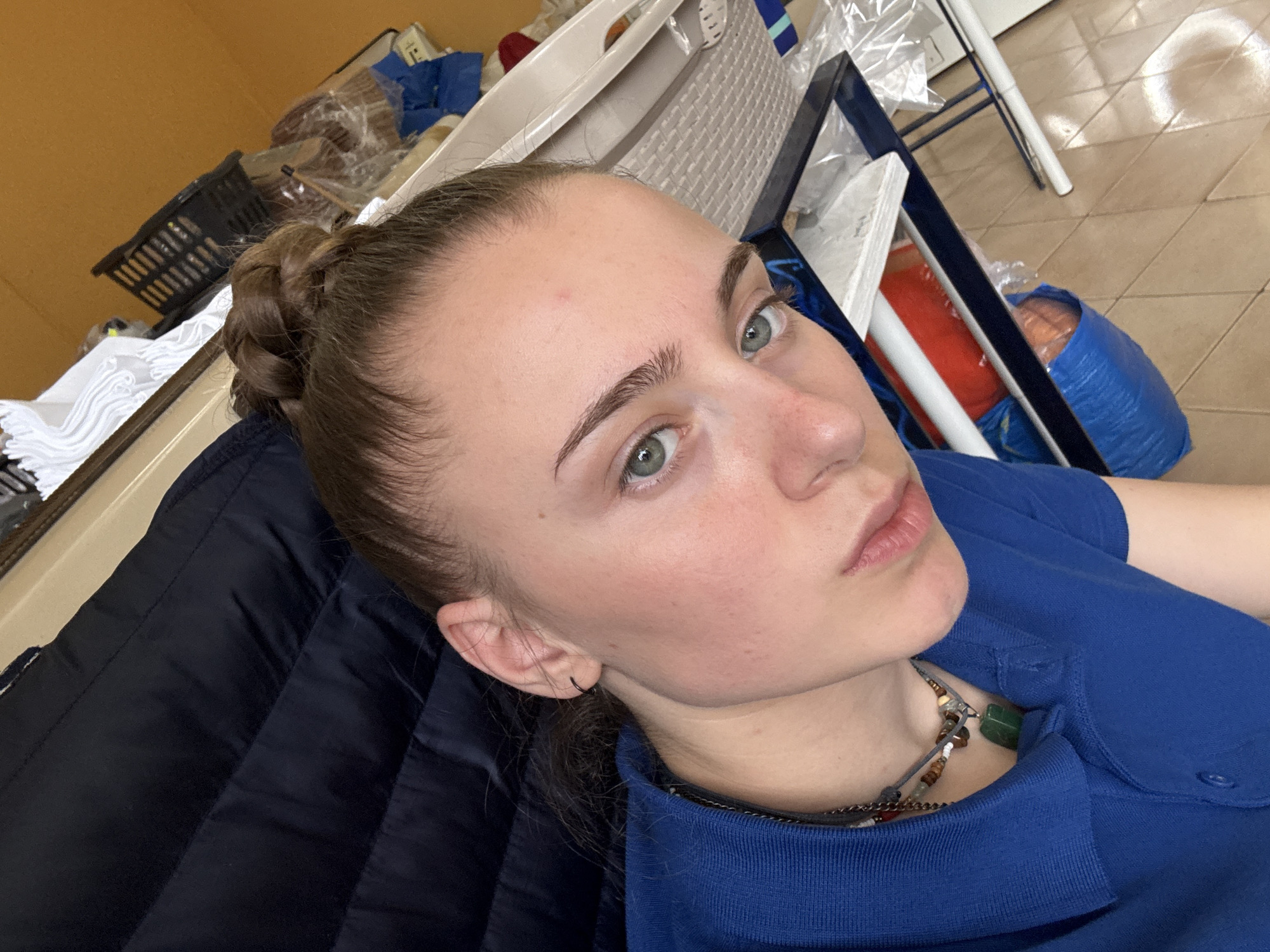 A close-up, angled shot of a young woman with light skin and green eyes. Her hair is pulled back in a bun at the crown of her head. She wears a blue polo shirt and a beaded necklace with a green pendant. Her left ear has a black hoop earring. She is leaning against a dark blue, quilted jacket. In the background, there is a cluttered space with a white plastic chair, a black basket, and various items wrapped in plastic. The tiled floor is visible. The specific location within Sofia, Bulgaria cannot be determined from the image.