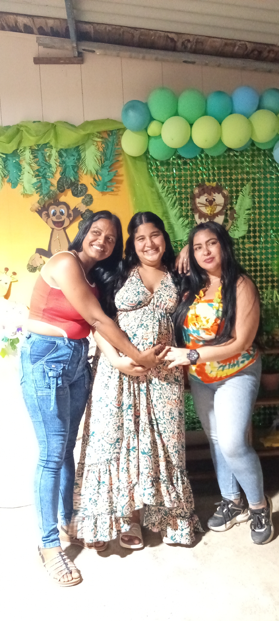Three women are posing for a photo in front of a decorated backdrop. The backdrop features jungle-themed decorations including green and yellow balloons, a monkey cutout, a lion cutout, and artificial green foliage.
The woman in the center is wearing a long, patterned dress. The woman on the left is wearing a red top and blue jeans with a unique pocket design. The woman on the right is wearing an orange and blue floral top and blue jeans. All three women are smiling and appear to be holding hands or touching each other's hands in the center of the frame.
The location appears to be indoors, possibly at a party or celebration.