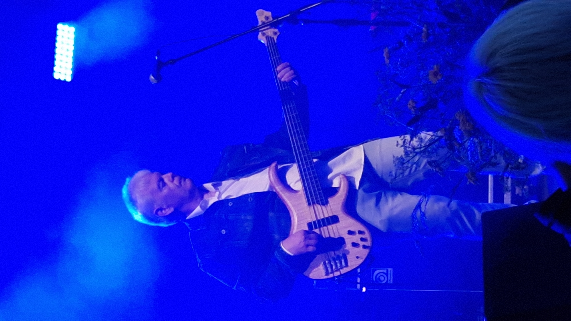 A man with gray hair plays a bass guitar on a dimly lit stage. He is wearing a dark jacket over a white shirt and light-colored jeans. A microphone stands to his right, and a bright light is visible in the background. The overall scene is bathed in a blue light, creating a smoky atmosphere. The image likely depicts a musical performance in Priekule, Latvia.