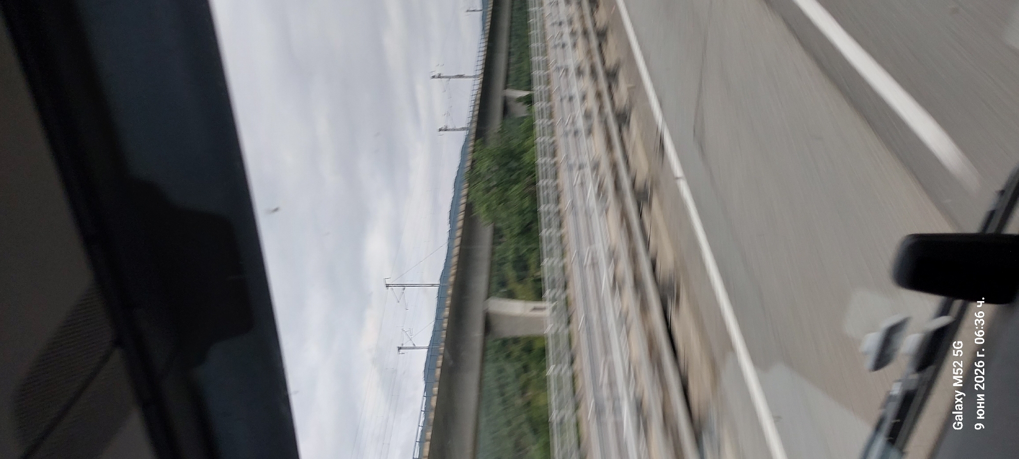 The image shows a view from a vehicle moving along a highway or elevated road. In the background, there is a large concrete overpass, likely carrying a railway line, supported by pillars and surrounded by green trees and hills under a cloudy sky. The foreground shows the road surface with white lane markings, and the interior of the vehicle is partially visible at the bottom, including a dashboard element and a phone displaying "Galaxy M52 5G" and the date "9 юни 2026 г. 06:36 ч." The scene is a perspective of transit through a landscape featuring infrastructure and natural elements. There are no people or specific interactions visible. The location is not explicitly indicated as La Jonquera, Spain within the image itself, but it could be a location near the border given the presence of highway infrastructure.