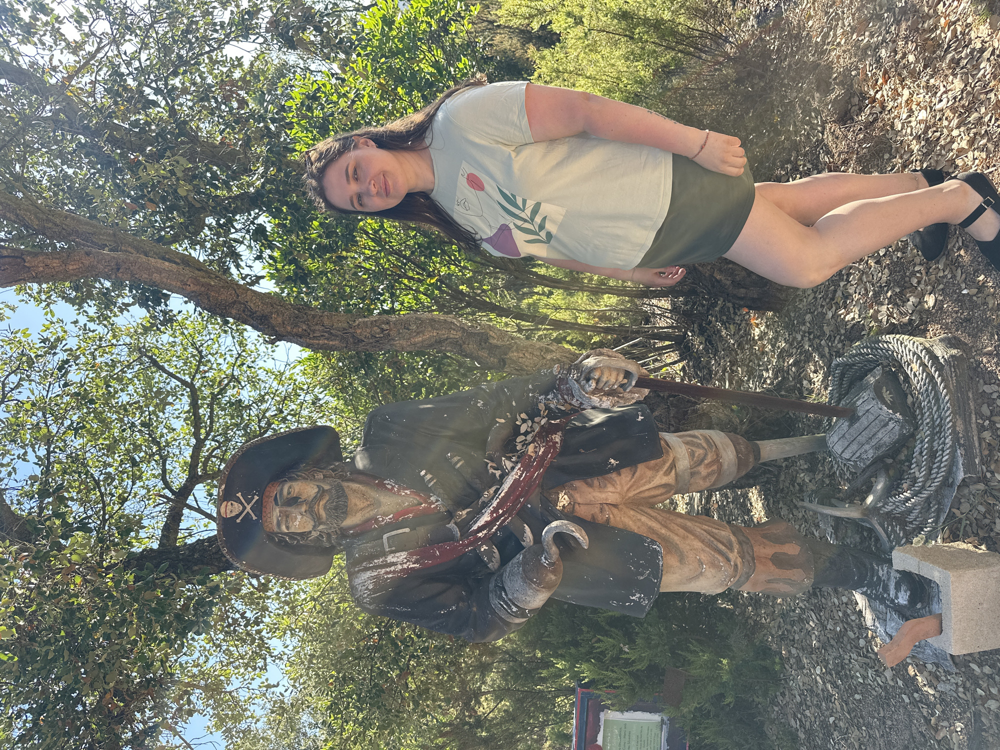 A young woman stands to the right of a weathered pirate statue in an outdoor setting, possibly a park or garden, with trees and foliage in the background. The woman is wearing a light green t-shirt with a graphic print of an abstract face and plant elements, and a dark green skirt. She has long brown hair and is looking towards the camera with a slight smile.
The pirate statue is a full-body sculpture, depicted wearing a large hat with a skull and crossbones emblem, a dark coat, a red sash, and light-colored pants. It has a hook for a left hand and appears to be holding a cane. The statue shows signs of age and weathering, with chipped paint and a worn appearance. At the base of the statue, there are nautical elements such as a coiled rope and an anchor.
In the background, lush green trees and bushes are visible, with sunlight filtering through the leaves. To the left of the pirate statue, there is a green sign with text on it, partially obscured by foliage. The ground is covered with leaves and dirt. The scene suggests a casual outdoor environment, potentially a tourist attraction or a themed area. The location in Sant Feliu de Guíxols, Spain, is not visually evident from the image itself.