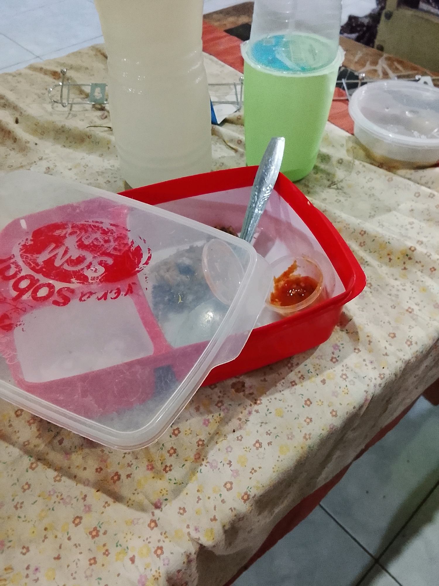 A close-up, slightly overhead view shows a red plastic food container on a floral patterned tablecloth. Inside the container, there are at least two compartments, one with a small plastic cup containing a red sauce. A spoon rests across the top of the container. To the left, the lid of the container is partially covering its contents, revealing a red circular logo with white lettering. Behind the container, a tall, translucent white bottle and a green plastic cup with a blue liquid are visible. A clear plastic container with a lid is also present to the right. The scene appears to be indoors, possibly on a table in Ujungrusi, Indonesia, given the context.
