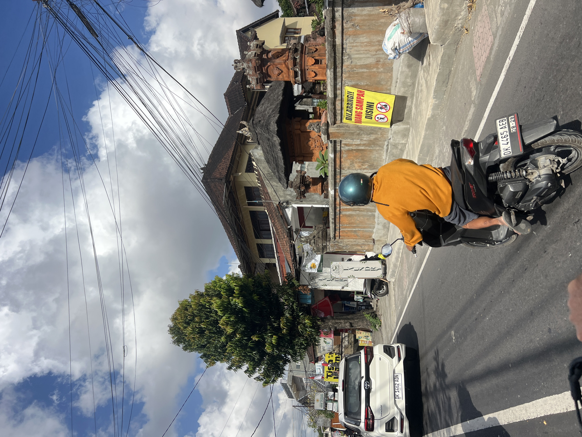 In Denpasar, Indonesia, a street scene unfolds under a partly cloudy sky with power lines crisscrossing overhead. A white SUV, with license plate DK 1462 ADN, travels on the left side of the road. In front of the SUV, a person wearing an orange shirt and a blue helmet rides a black scooter, their license plate reading DK 4465 AEB. Adjacent to the road, a concrete wall displays a yellow sign with red text that reads "DILARANG!! BUANG SAMPAH DISINI," accompanied by pictograms of people disposing of trash. Beside the wall, a large white plastic bag filled with waste rests on the ground. Behind the wall, a traditional Balinese structure with intricate stone carvings and a thatched roof is visible, alongside a multi-story building. A tall, leafy tree stands prominently near the road, with various shops and signs lining the street.