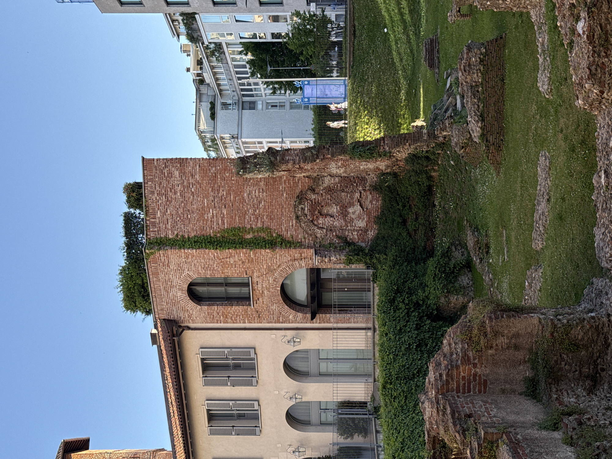 The image shows a vertical composition featuring a combination of architectural elements and greenery. On the left, there is a building with a beige facade, distinguished by rows of shuttered windows and arched windows on the upper levels. Adjacent to it is a brick structure that rises higher, adorned with vines and plants growing on its surface.
On the right, the ruins of the Roman amphitheater of Milan, now a historical site overgrown with grass and vegetation, are visible. In the background, modern buildings can be seen, including a building with multiple floors and windows, suggesting an urban setting. The sky is clear and blue, indicating daylight conditions. The scene captures a juxtaposition of ancient ruins and modern urban architecture in Milan, Italy.