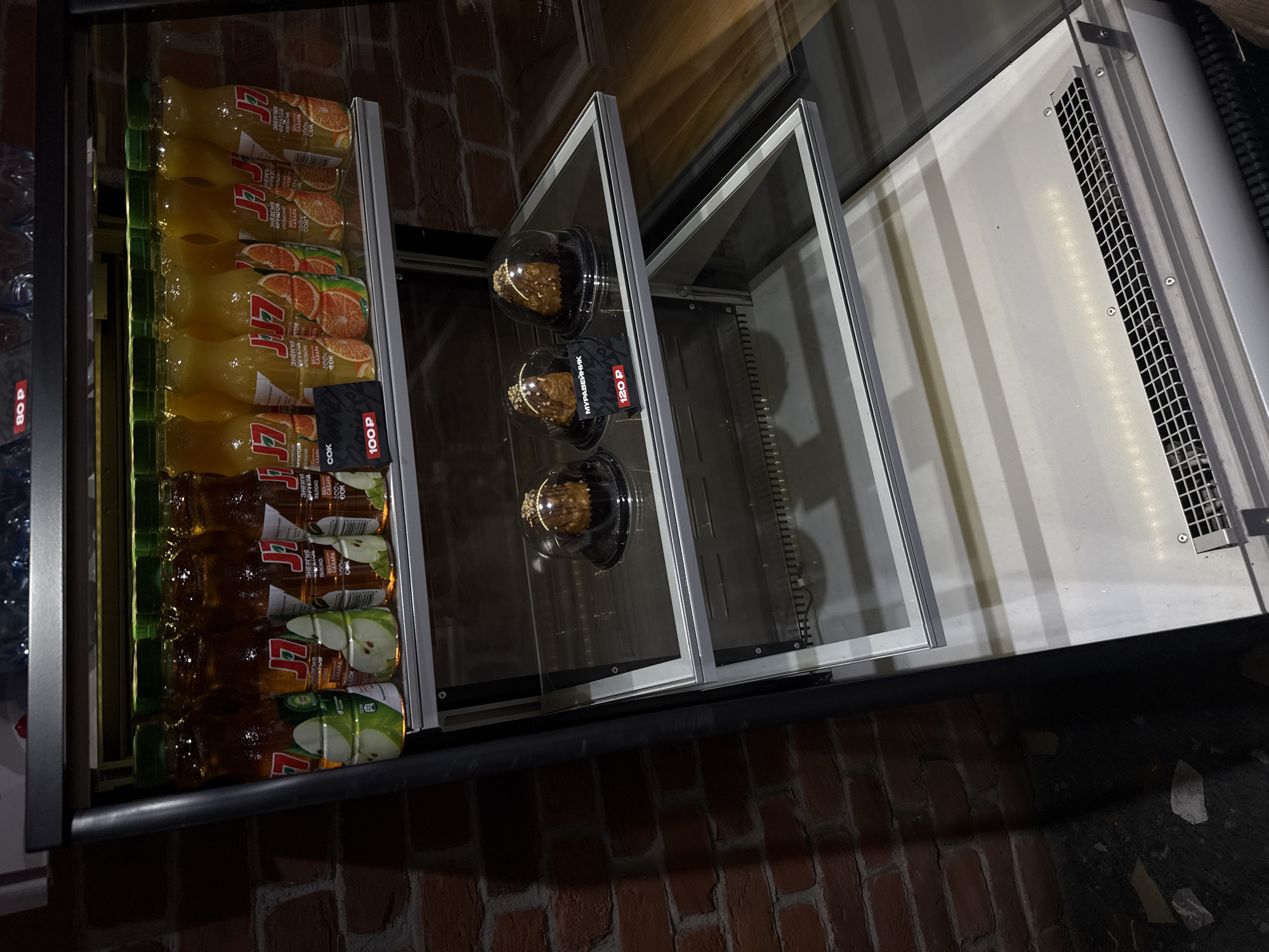 An indoor, vertically oriented shot displays a refrigerated display case filled with beverages and desserts, suggesting a convenience store, cafe, or similar food establishment. The background features a reddish-brown brick wall, while the floor appears dark and uneven, possibly concrete or pavement.
On the left side of the display, numerous plastic bottles of juice are neatly stacked in rows. The bottles are primarily yellow-orange and brown, indicating different flavors. Visible text on the labels includes "СОК" (Juice), "ЭНЕРГИЯ ФРУКТОВ" (Energy of Fruits), "АПЕЛЬСИН" (Orange), and "ЯБЛОКО" (Apple), with some labels also mentioning "100%" and "БЕЗ САХАРА" (No Sugar) or "CAXAPA" (Sugar). A brand logo, "7Д", is also prominent. Price tags are attached to some of the juice bottles, showing "80 P" and "100 P".
To the right of the juice bottles, within clear, modular compartments of the display, three individual desserts are visible. Each dessert is encased in a clear plastic dome. One of the desserts has a black price tag with white text that reads "МУРАВЕЙНИК" (Muraveynik, a type of Russian cake) and "120 P". The desserts appear to be a type of pastry or cake, possibly with a crumbly texture and nuts.
The rightmost section of the display case is partially empty, showing metallic shelving and a white interior with a rectangular grate, likely part of the cooling mechanism. The overall lighting is artificial and casts some shadows. The image is taken from a slightly elevated perspective looking down into the display.