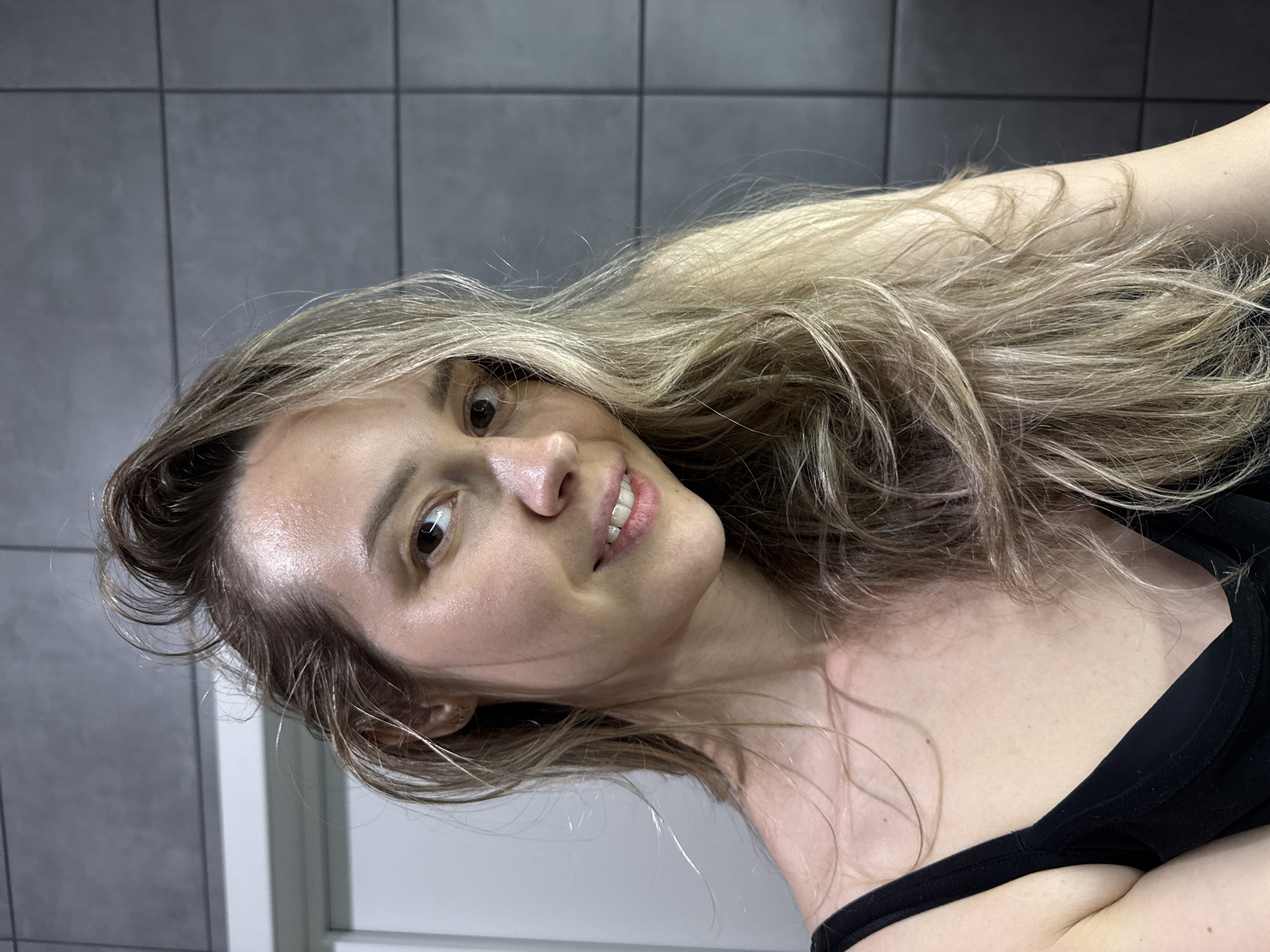 A headshot of a young woman is presented, with the original image rotated ninety degrees counter-clockwise. The woman's head is tilted slightly, and she looks directly at the viewer with open, brown eyes and a subtle, natural smile, showing a hint of her upper teeth. Her long, wavy hair, a mix of blonde and light brown, flows around her shoulders and chest. Her complexion appears clear and slightly dewy, with highlights visible on her forehead and cheekbones. She is wearing a black top, with a strap visible over her right shoulder. The background consists of a gray tiled wall with dark grout lines, suggesting an indoor, possibly utilitarian setting like a bathroom or hallway. The lighting is bright and direct. No specific activity is depicted other than posing for the photograph. There is no visible text in the image. The location context is Chaikovsky, Russia.