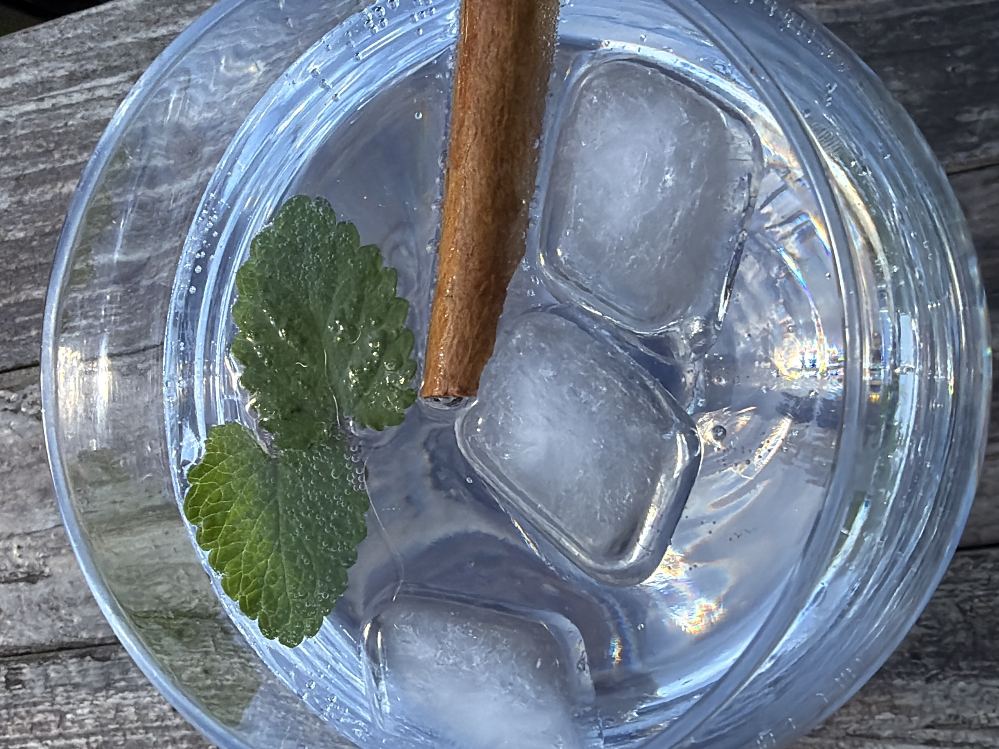 A top-down close-up shot depicts a clear glass filled with liquid, ice cubes, a cinnamon stick, and a mint leaf. The glass sits on a weathered wooden surface. Esch-sur-Alzette, Luxembourg is not discernible from the image.