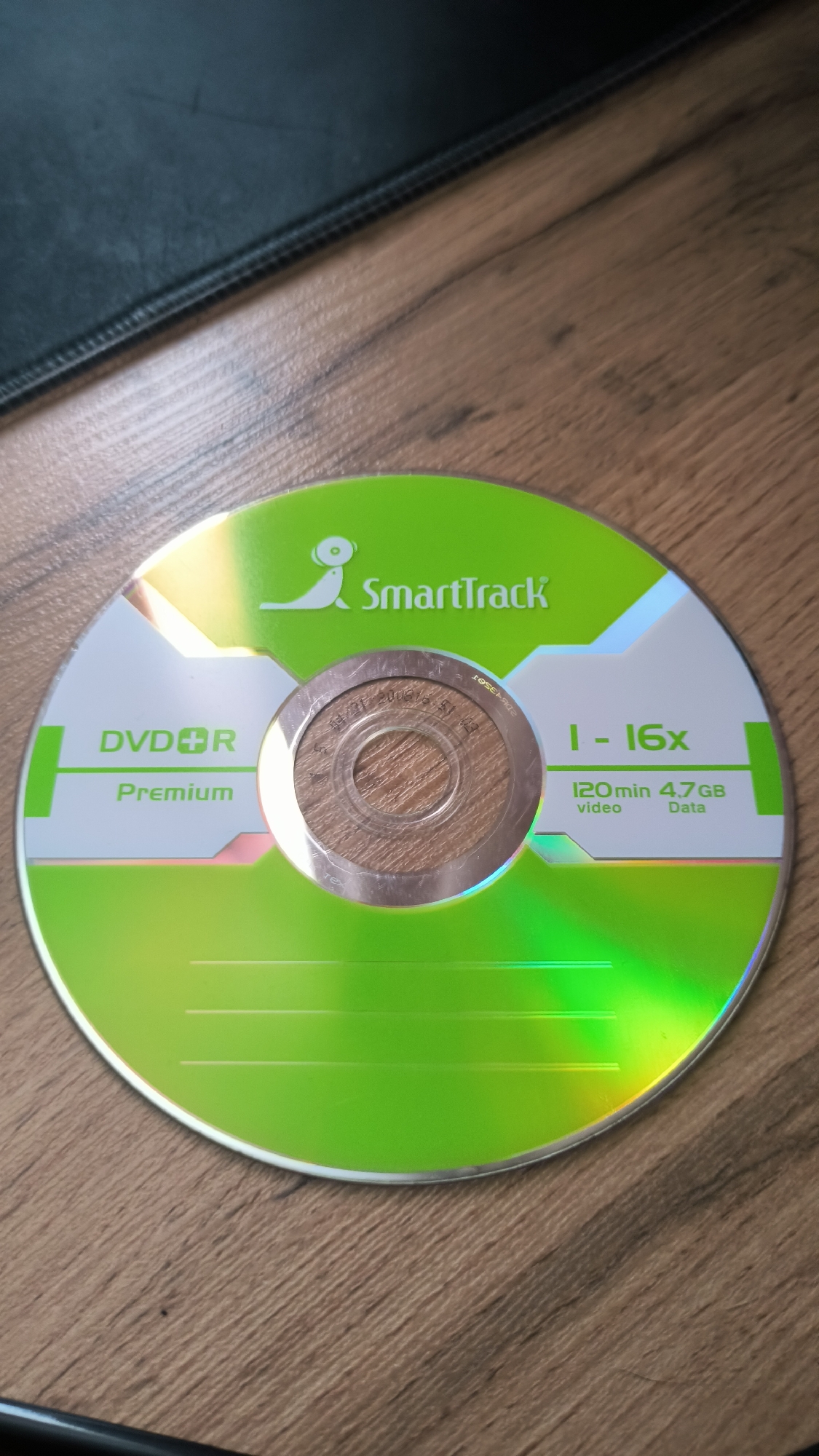The image shows a DVD-R disc with a bright green and white design. The disc is lying on a wooden surface, likely a table, with a dark, textured object, possibly a mousepad or a book, in the upper left corner. The disc itself has several lines of text and graphics.
In the center of the disc, there's a silver hub with some faint, etched text that is difficult to read. To the left of the hub, the text "DVDR" and below it "Premium" are prominently displayed in white lettering.
To the right of the hub, there are green text elements indicating "1 - 16x". Below this, "120 min" and "4.7GB" are listed, with "video" and "Data" beneath them respectively. A stylized logo featuring what appears to be a leaping figure or animal is positioned above the text "SmartTrack" in green lettering. The overall design is sleek and modern, with the green and white colors dominating the disc's surface. The disc is reflecting light, creating a rainbow effect, suggesting it is under direct lighting. The scene appears to be indoors, given the lighting and the wooden surface. No people or specific events are depicted. The location context of Kovrov, Russia, is not visually evident from the image itself.