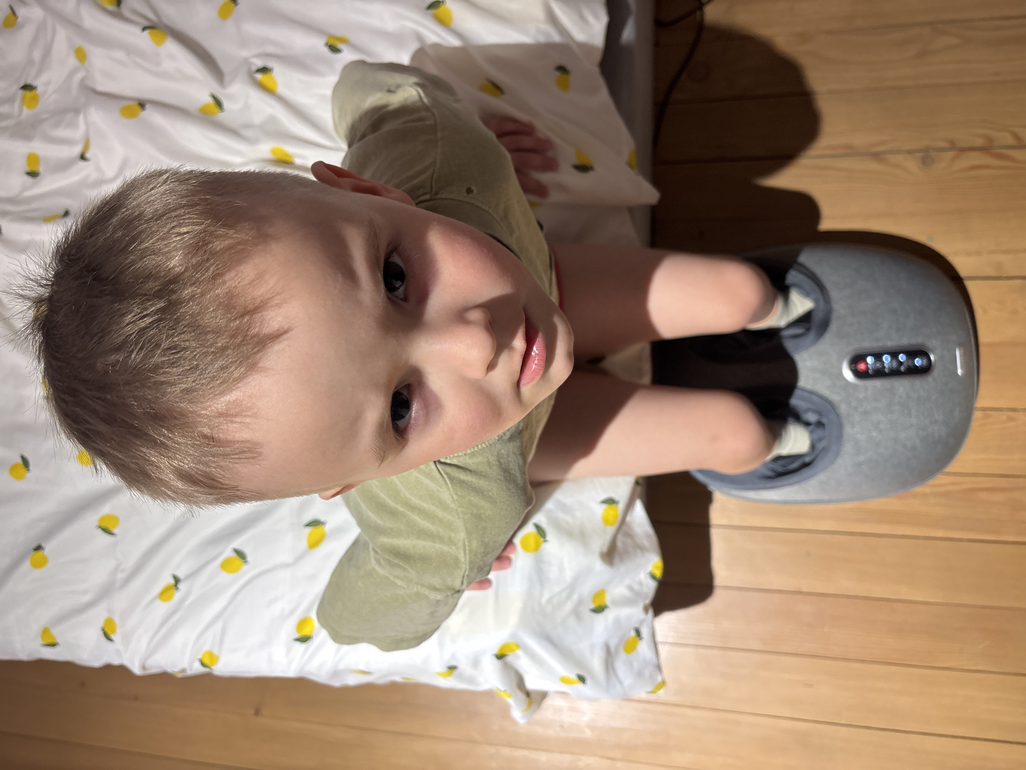 A young child stands on a wooden floor, their feet inside a gray foot massager. The child is wearing a green shirt, and their arms are crossed. A white sheet with a lemon pattern is visible in the background. The setting appears to be indoors, possibly a home. There is no location context of Chaikovsky, Russia.