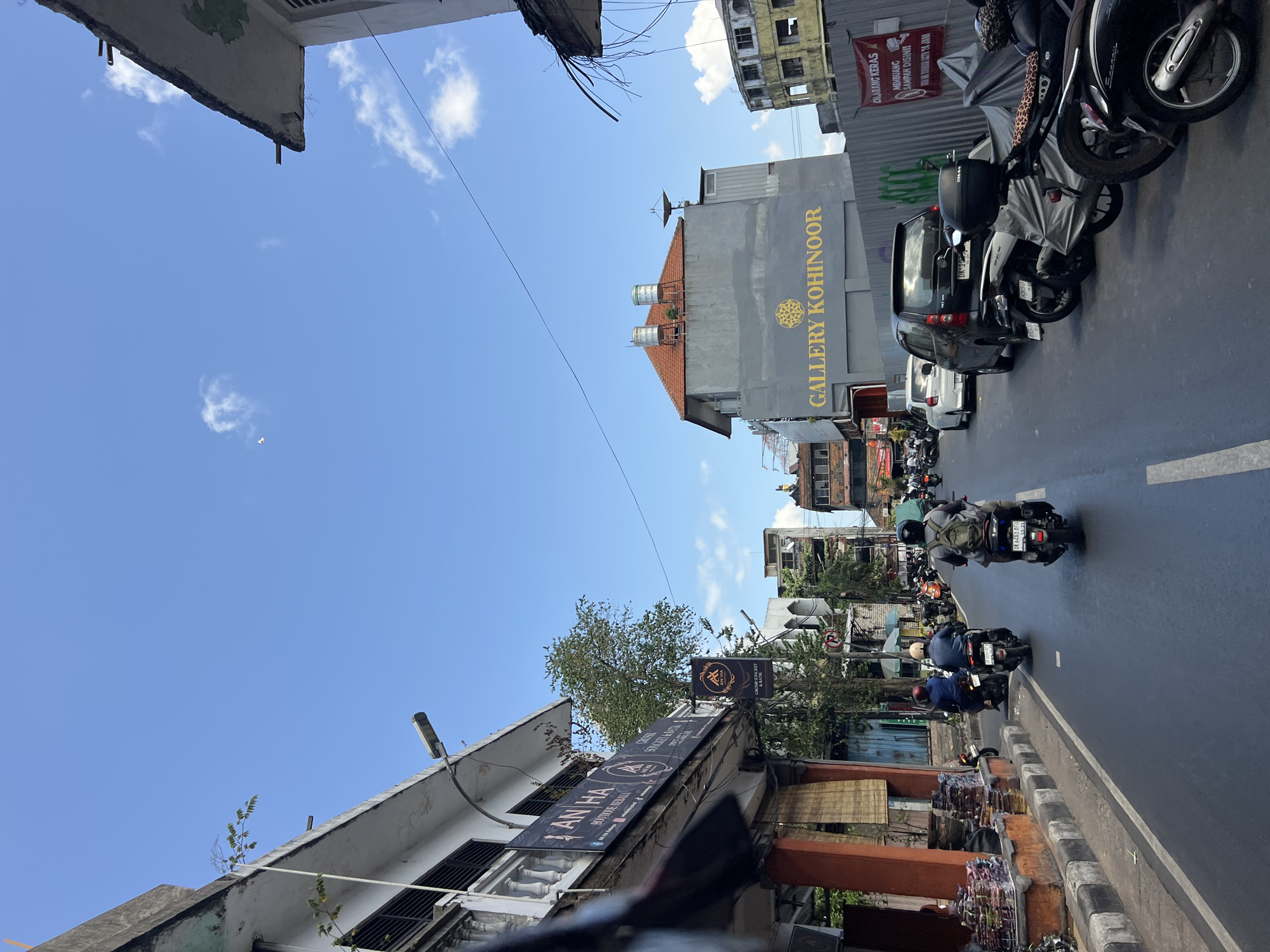 In Denpasar, Indonesia, a street scene unfolds under a bright blue sky with scattered clouds. On the left side of the road, shops line the sidewalk, one displaying banners for "AN HA" which appears to be a boutique selling clothing, possibly kebaya and batik. Merchandise is stacked on the sidewalk in front of another shop. The road itself is busy with traffic, primarily motorcycles. Several individuals are riding motorcycles in the same direction, their vehicles filling the lanes. Parked motorcycles are visible along the right side of the road. Beyond the shops and parked vehicles, a building with the text "GALLERY KOHINOOR" is prominent. Further down the street, more buildings are visible, some appearing to be under construction or in disrepair. A red banner with white text is attached to a fence, containing a warning in Indonesian. The overall impression is of a bustling urban environment with active commerce and transportation.