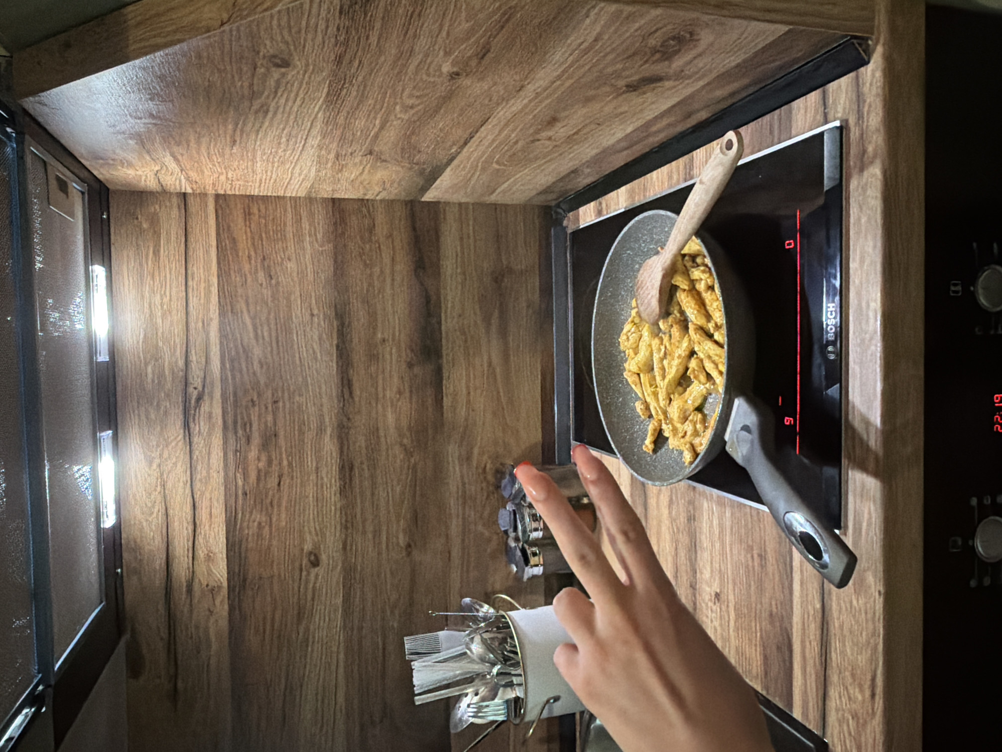 In a kitchen, a hand with polished fingernails forms a peace sign in front of a frying pan on a stovetop. The pan contains a portion of golden-brown pasta with sauce. A wooden spoon rests inside the pan, and the stovetop displays red indicator lights. The background features wooden paneling and cabinetry. The kitchen appears to be part of a residential dwelling located in Varna, Bulgaria.