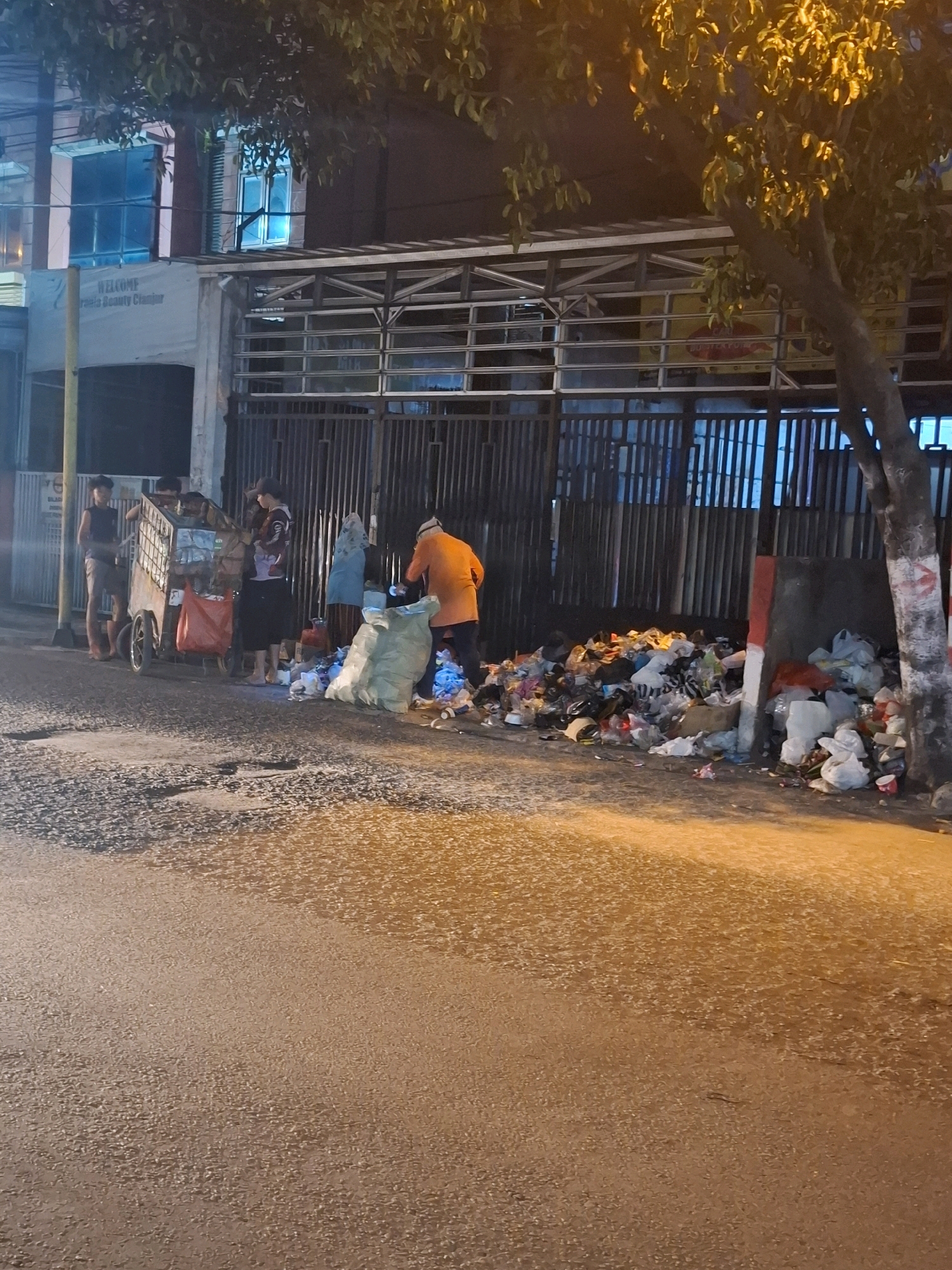 In Cianjur, Indonesia, a street scene unfolds at night with a substantial accumulation of garbage on the roadside. A metal fence forms a backdrop to the scene, behind which a building with windows illuminated by blue light is visible, bearing the "WELCOME" signage for "Gisela Beauty Center." Several individuals are present near a three-wheeled cart laden with items, some of which appear to be trash bags. One person, wearing an orange shirt, is actively engaged with the garbage pile, and two large, light-colored bags are placed beside the heap. The street itself is paved and uneven, reflecting the ambient light. A tree with illuminated leaves stands to the right of the frame, partially obscuring the view.