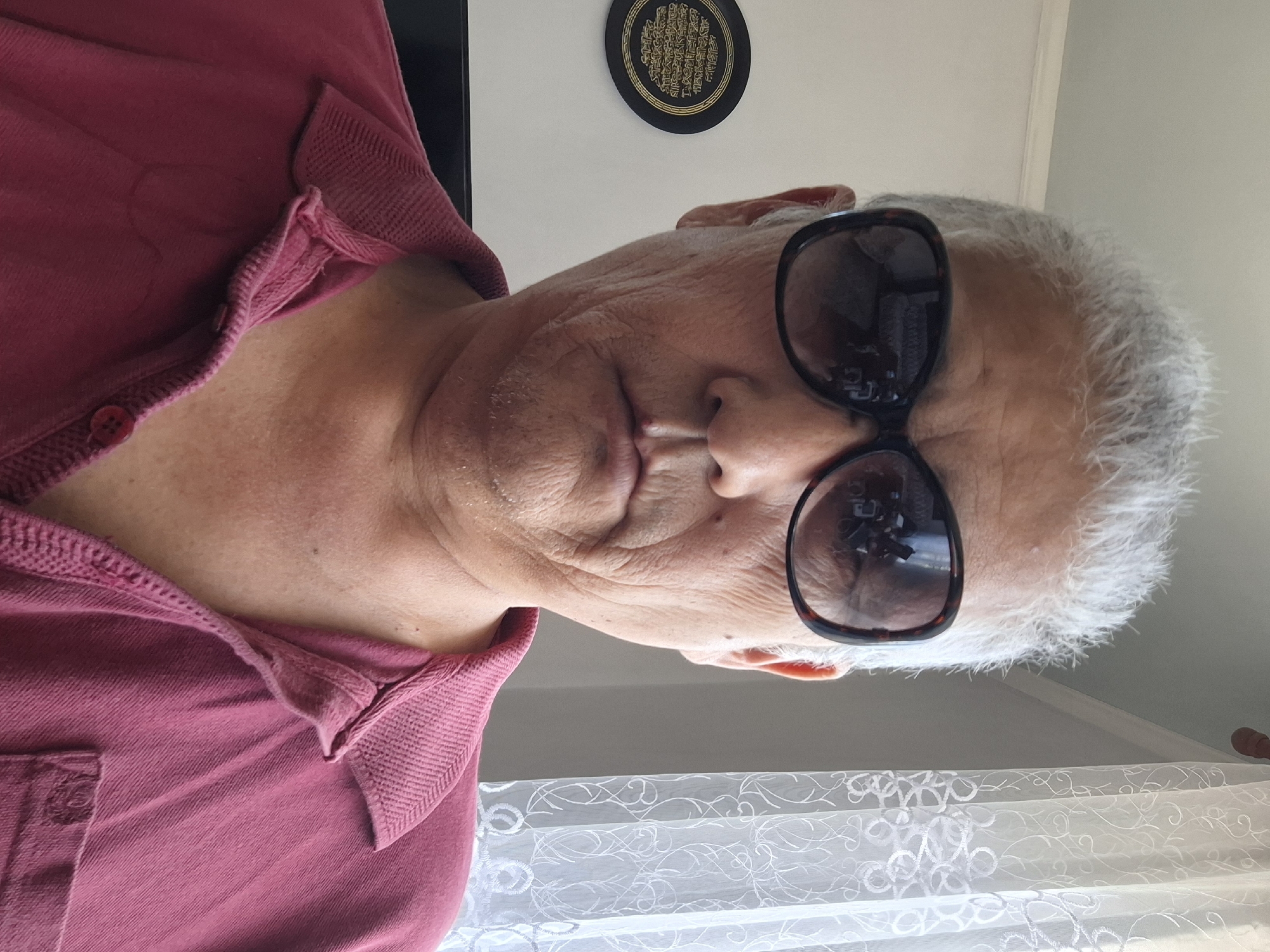 A close-up, head-and-shoulders selfie of a person wearing large, dark-rimmed sunglasses and a maroon collared shirt. The person has gray hair and visible wrinkles on their face. In the background, on the left, is a circular black and gold decorative plate on a white wall. To the right, a white lace curtain is visible. The lighting suggests an indoor setting during the daytime. There is no visible indication of the specific location within Almaty, Kazakhstan.
